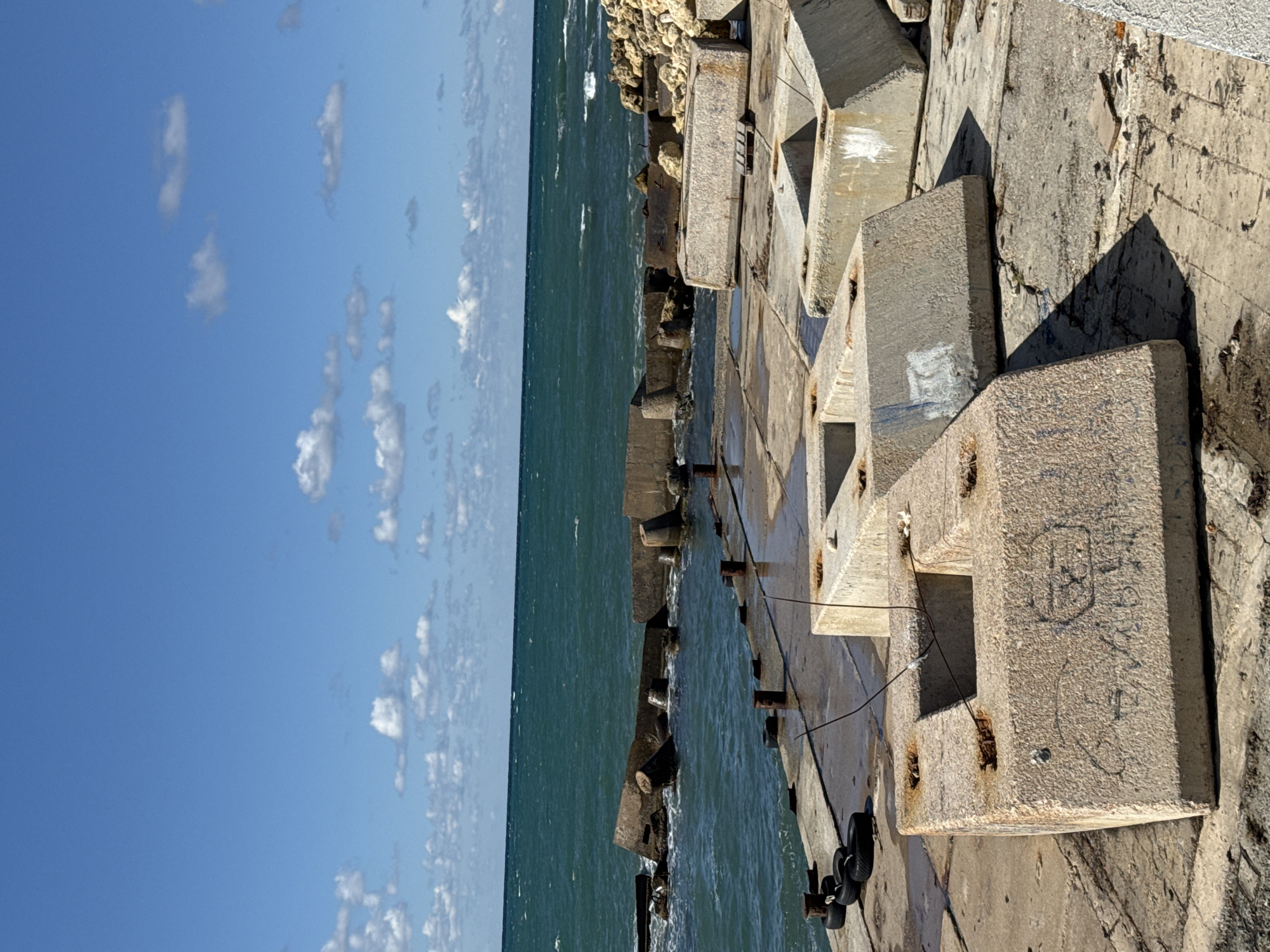 The image shows a concrete pier with various concrete blocks scattered on it, some in the foreground and others extending towards the sea. The sea is a deep teal color with white waves breaking near the shore. In the background, there are remnants of a breakwater made of concrete structures and rocks. The sky is a bright blue with scattered white clouds. There are no people visible in the image. The scene appears to be a coastal area, possibly a harbor or a protected shoreline, with evidence of construction or erosion control measures. The overall impression is one of a utilitarian and somewhat weathered marine environment.
