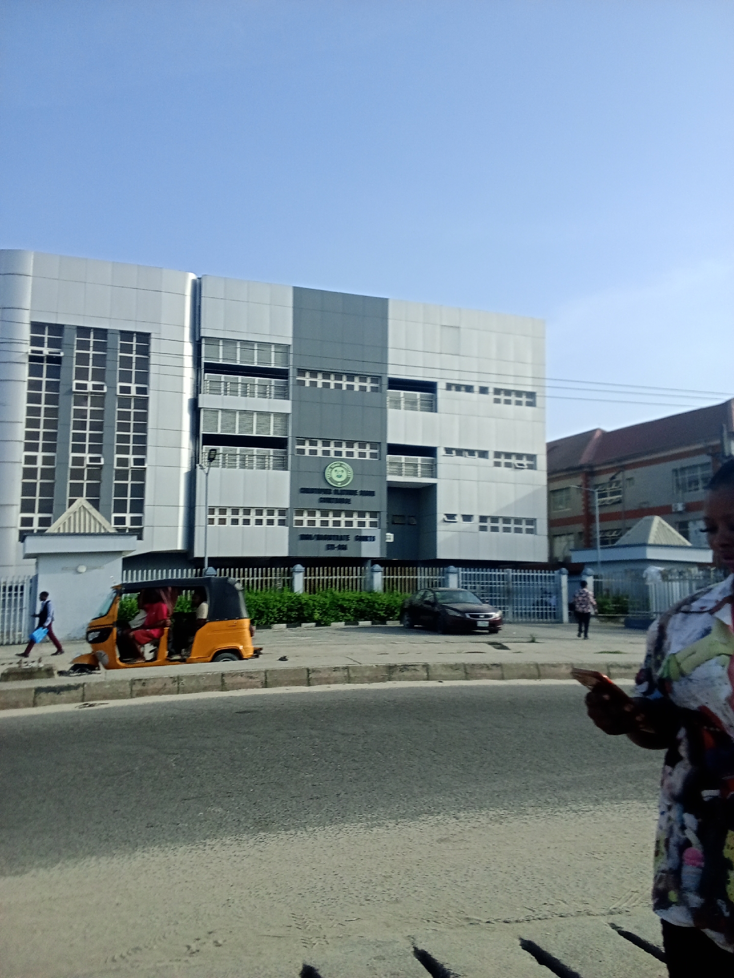 The image shows a street scene in Ado, Nigeria. A modern, multi-story building with a gray and white facade is visible in the background. The building appears to be a courthouse based on the inscription "SHARIA LAW COURTS COURT COMPLEX MAGISTRATE COURTS 173-402". A yellow auto rickshaw (tuk-tuk) is parked near the sidewalk in front of the courthouse. A dark colored sedan is also parked nearby. Several people are present: one man walks to the left of the tuk-tuk, and a woman is standing on the sidewalk to the right, partially obscured by the camera. The sky is clear and blue.