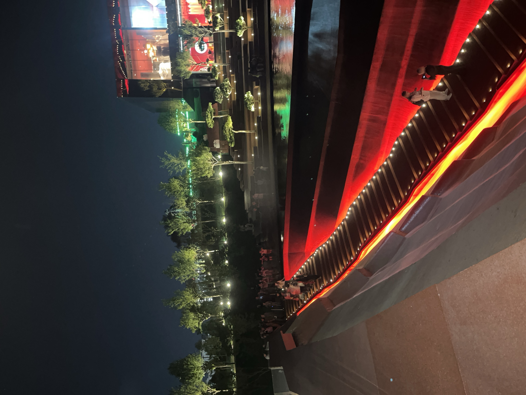 The image depicts an outdoor nighttime scene, likely in Krasnodar, Russia. The setting appears to be a public space with a modern architectural design, possibly a promenade or a plaza. A prominent feature is a long, tiered structure with red and yellow lighting, creating a vibrant and dramatic effect. This structure seems to be a bridge or a raised walkway, lined with small, bright lights along its edge.
Several people are visible on and around this illuminated structure. Some are standing and observing, while others are walking. Two individuals are particularly noticeable: one dressed in white, possibly a dancer or performer, and another in dark clothing. Their presence suggests an event or a planned gathering is taking place.
To the left of the illuminated structure, a line of trees with scattered lights can be seen. These trees are illuminated from below, casting a soft glow and adding to the festive atmosphere. In the background, a large building with illuminated windows is visible, suggesting commercial or residential activity. The overall impression is one of a lively evening event in an urban environment, characterized by striking lighting and a gathering of people. The dark sky indicates it is nighttime. There is no visible text in the image.