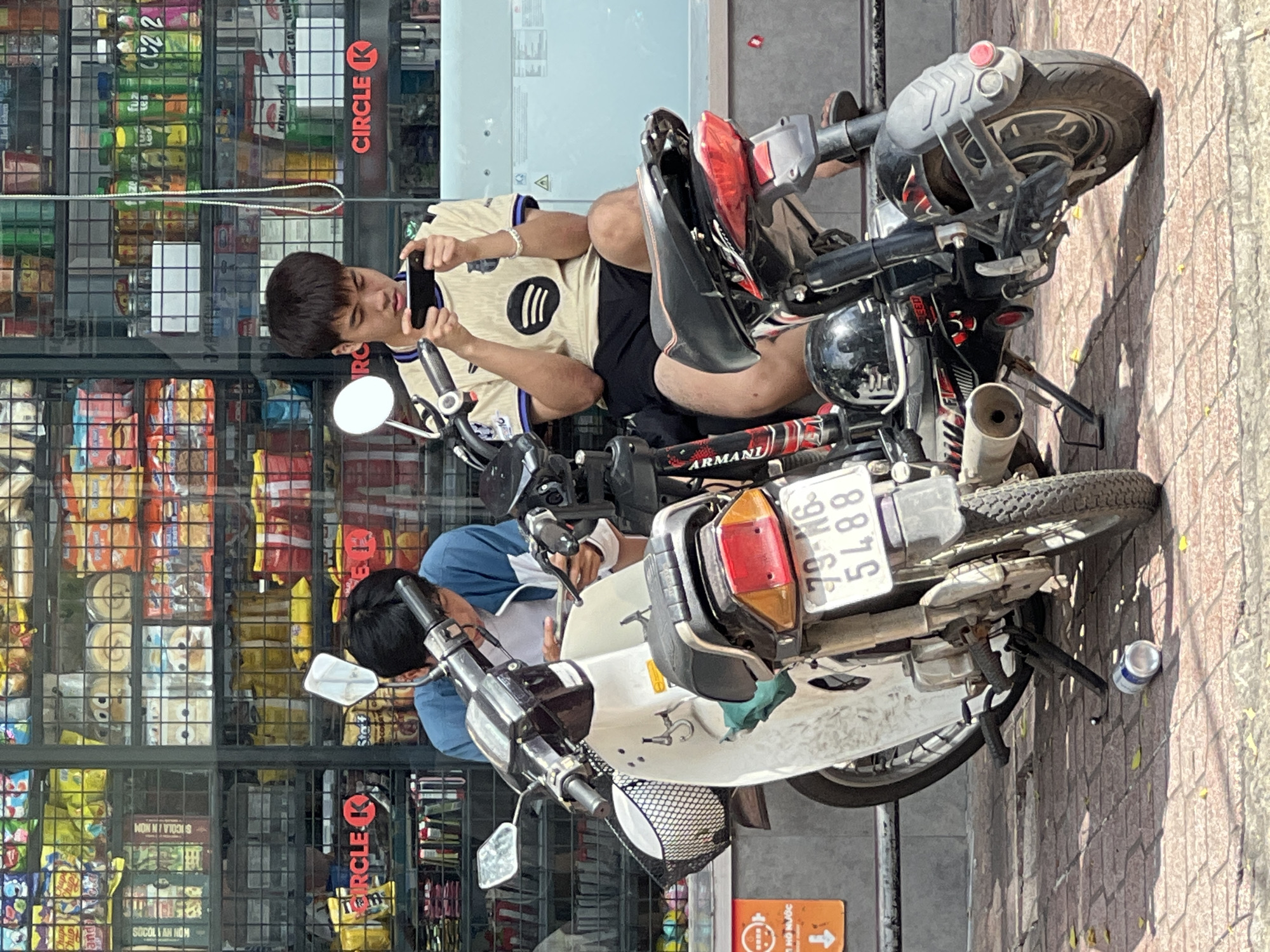 In the city of Nha Trang, Vietnam, two young men are seated on motorcycles parked on a brick sidewalk in front of a brightly lit convenience store. The store's shelves are stocked with a variety of products, including snacks and drinks. The man on the left, wearing a blue and white jacket, is looking down at his phone. The man on the right, in a yellow and black shirt with a Spotify logo, is also engrossed in his phone, holding it horizontally. Both men are casually dressed and appear relaxed. The motorcycles are parked close together, with one in front of the other. A discarded aluminum can lies on the sidewalk near the front wheel of the motorcycle on the left. The scene is illuminated by daylight, casting shadows on the ground.