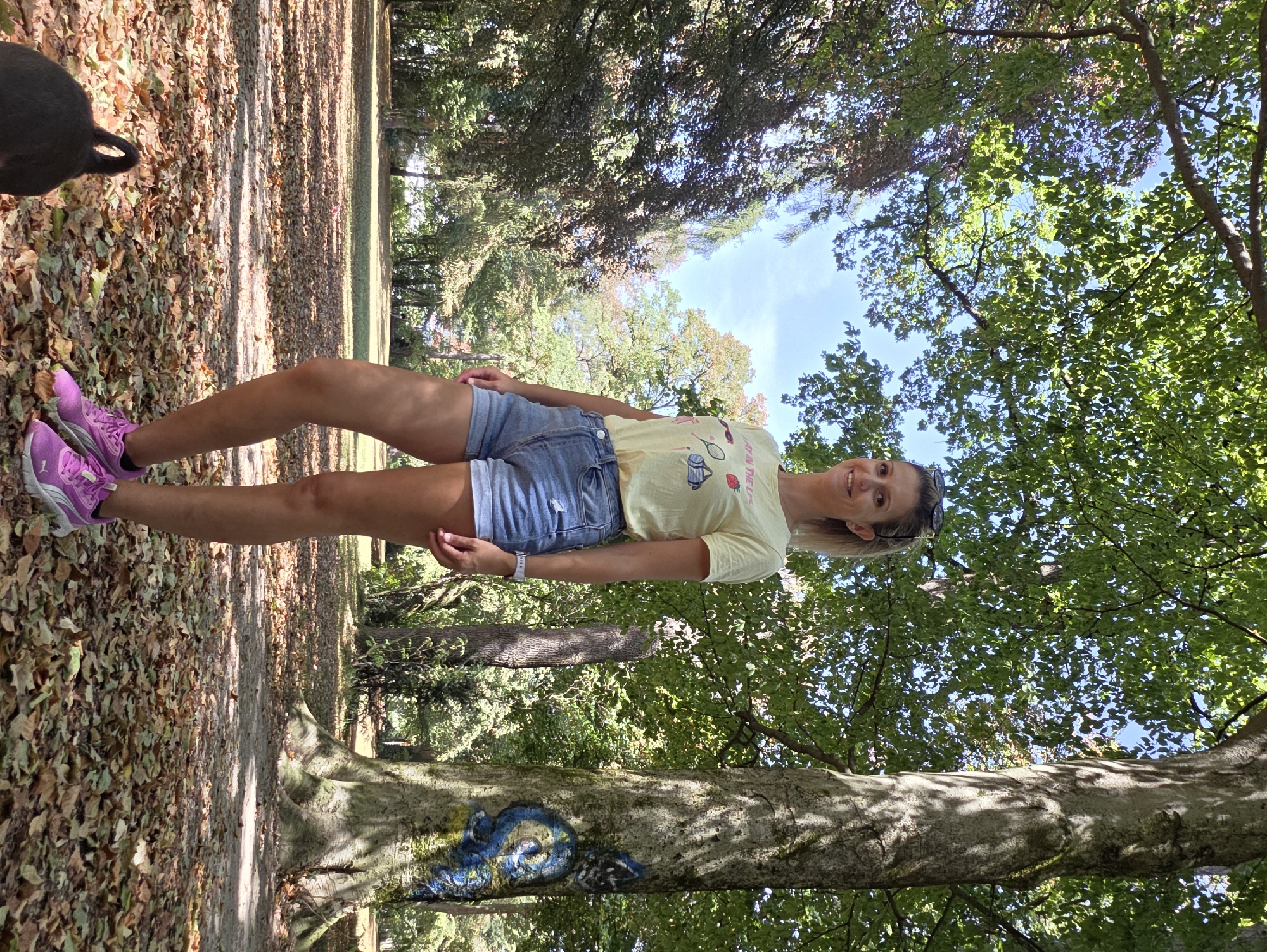 A blonde woman stands in a park on a sunny day. She is wearing a yellow t-shirt with a graphic print, denim shorts, and pink athletic shoes. She is smiling and looking at the camera. The park has a large tree with graffiti on its trunk to her right. The ground is covered with fallen leaves. A dark-colored object, possibly a dog, is visible in the lower left corner of the image. The background shows more trees and a clear blue sky. The scene appears to be the Englischer Garten in Munich, Germany.