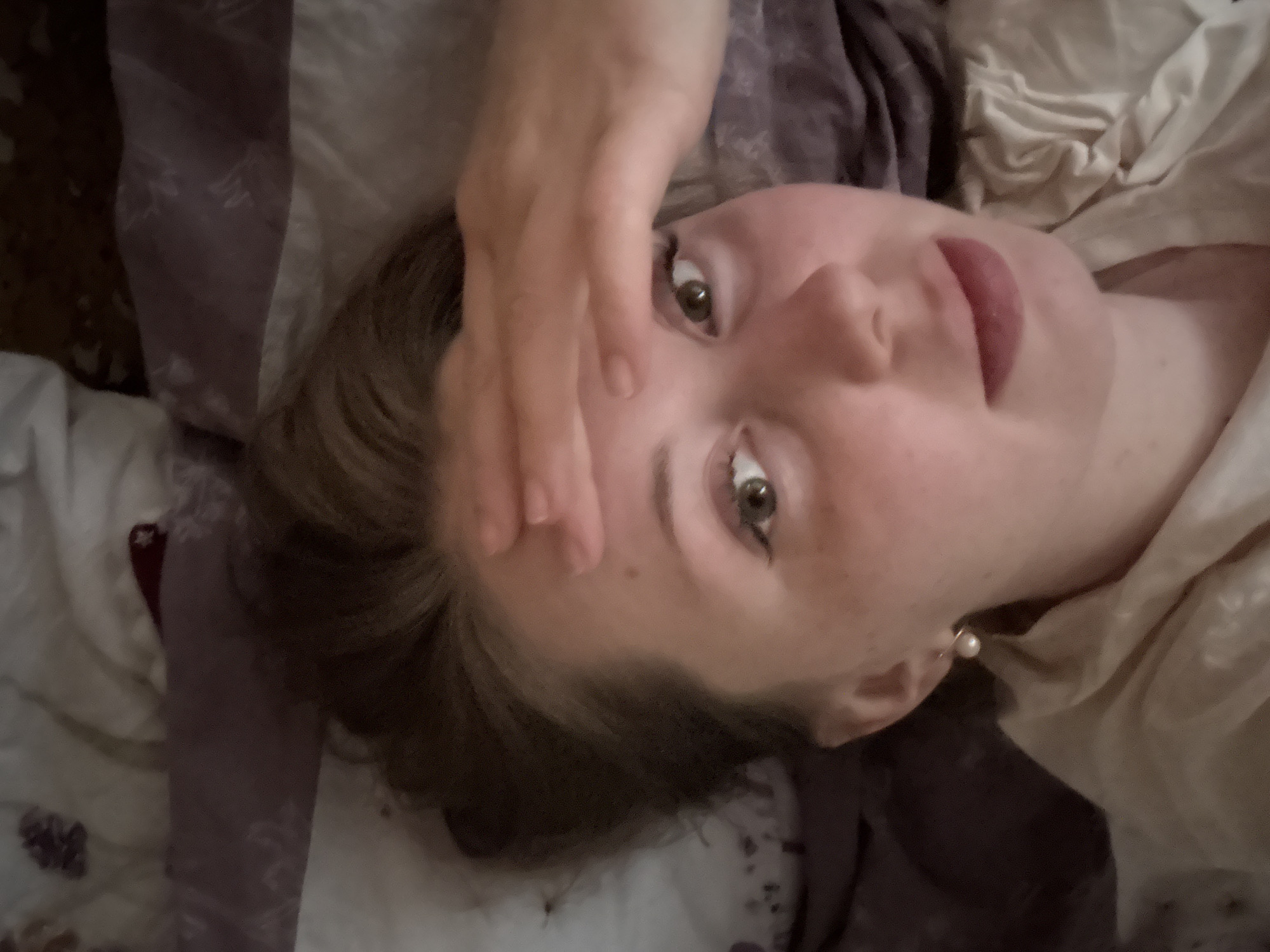 The image features a young person, appearing female, lying down and looking directly at the camera. Her head is slightly tilted, and her expression suggests a soft, subtle smile. She has light-colored skin, brown hair, and striking green or hazel eyes.
Her right hand is raised, fingers splayed, partially covering her forehead and the upper part of her face, including one eye. This gesture could imply shyness, shielding her eyes from light, or a relaxed, casual pose. She wears a small, spherical pearl earring in her visible left ear. Her lips have a natural, pinkish hue.
She appears to be dressed in light-colored, possibly off-white or cream, soft fabric around her neck and shoulder area. She is lying on what seems to be a bed, surrounded by soft, rumpled bedding. The bedding includes patches of white/cream and a darker, possibly patterned, purple or grey fabric. The scene suggests an intimate, indoor setting, likely a bedroom. The lighting is soft and diffused. There is no visible text in the image.