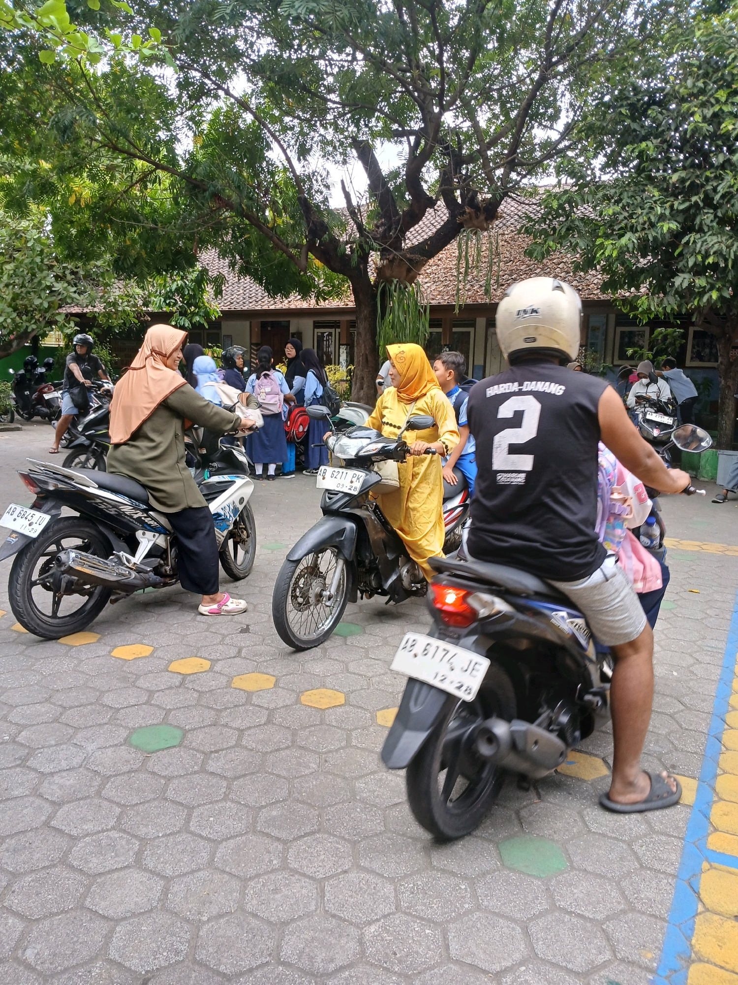 In Depok, Indonesia, on a paved hexagonal stone surface, a group of people are gathered around motorcycles. A woman wearing a tan hijab and olive-green tunic, with black pants and pink slip-on shoes, is seated on a white and blue motorcycle. To her right, another woman in a yellow hijab and long yellow dress is also on a motorcycle, holding the handlebars. In the foreground, a person wearing a white helmet and a black sleeveless shirt with the number "2" and text "HARDA-DANANG" on the back is on a blue and black motorcycle, facing away from the camera. This person is wearing light gray shorts and dark gray flip-flops. Several other motorcycles are visible, and a building with a tiled roof and some trees are in the background. The scene appears to be a pickup or drop-off point, possibly near a school or educational institution, given the presence of younger individuals in uniforms in the background.