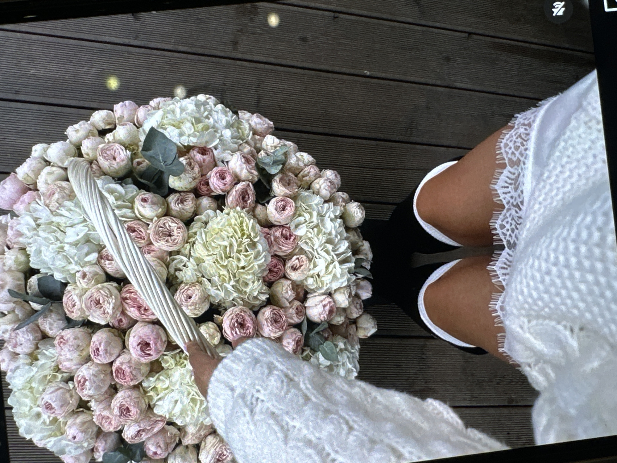 A person holds a basket filled with a large bouquet of light pink and white flowers. The person wears a white sweater and a white dress with a lace hem, paired with black knee-high socks with white trim. They stand on dark wooden planks, possibly a deck or walkway. The location cannot be determined.