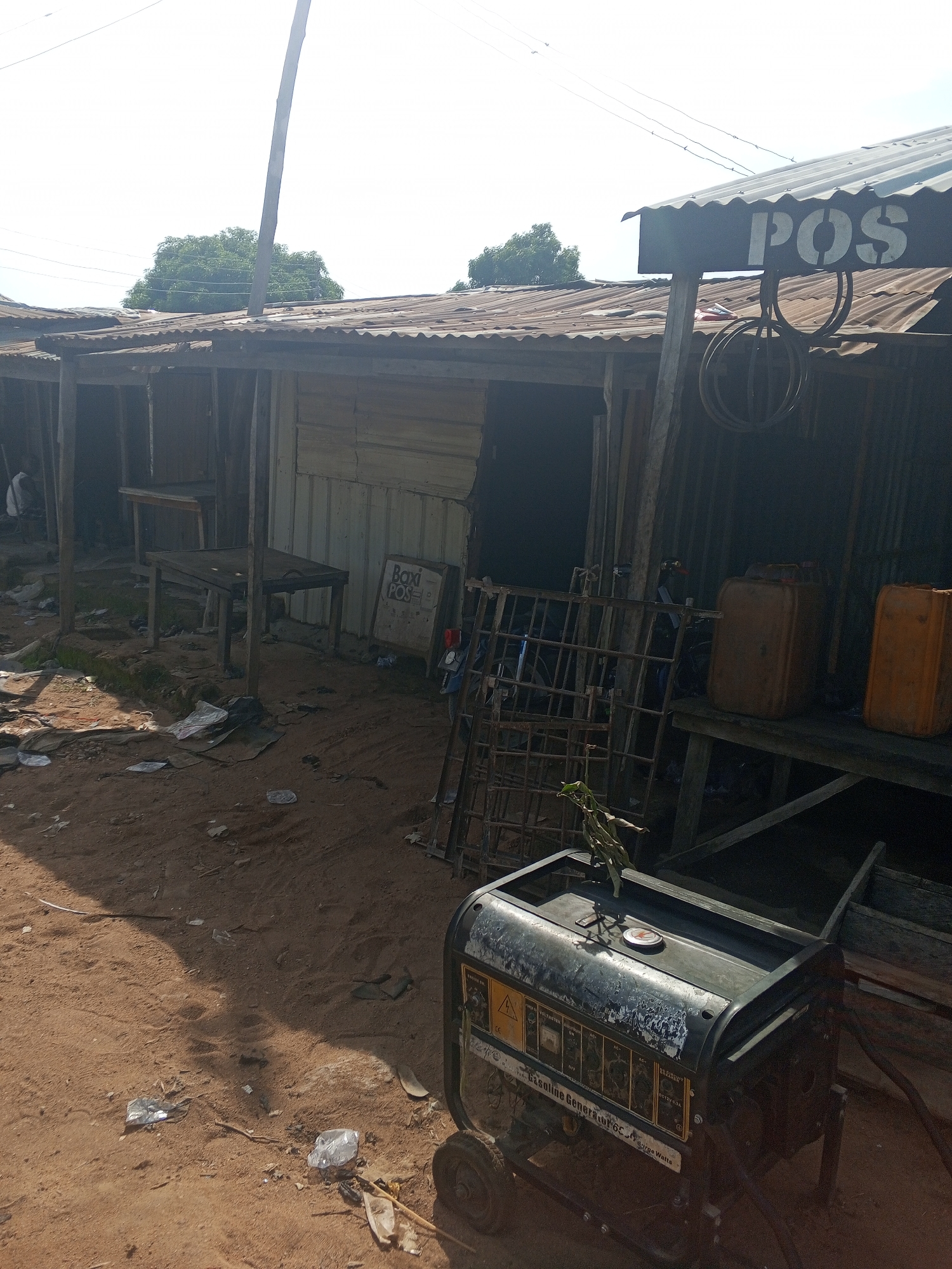 In Jalingo, Nigeria, an outdoor scene depicts a collection of structures with corrugated metal roofing, suggesting a commercial or makeshift market area. In the foreground, a dark-colored gasoline generator with visible wear and tear sits on the sandy ground. A label on the generator indicates "GASOLINE GENERATOR" and "Surge Watts." To the right of the generator, a wooden shelf holds two plastic jerrycans, one orange and one a darker, possibly brown, color. Behind these, a metal grate structure is visible. Further back, several open-fronted stalls or shops are evident, constructed from wood and metal sheeting. One stall has a sign with "POS" painted in large white letters. Inside this stall, two wooden tables are positioned. Another stall displays a sign for "Baxi POS". The ground is covered in dirt and scattered debris, including plastic waste. Overhead, power lines are strung across the sky, and trees are visible in the background. The overall impression is one of a functional, albeit somewhat unkempt, commercial hub in an urban or semi-urban setting.