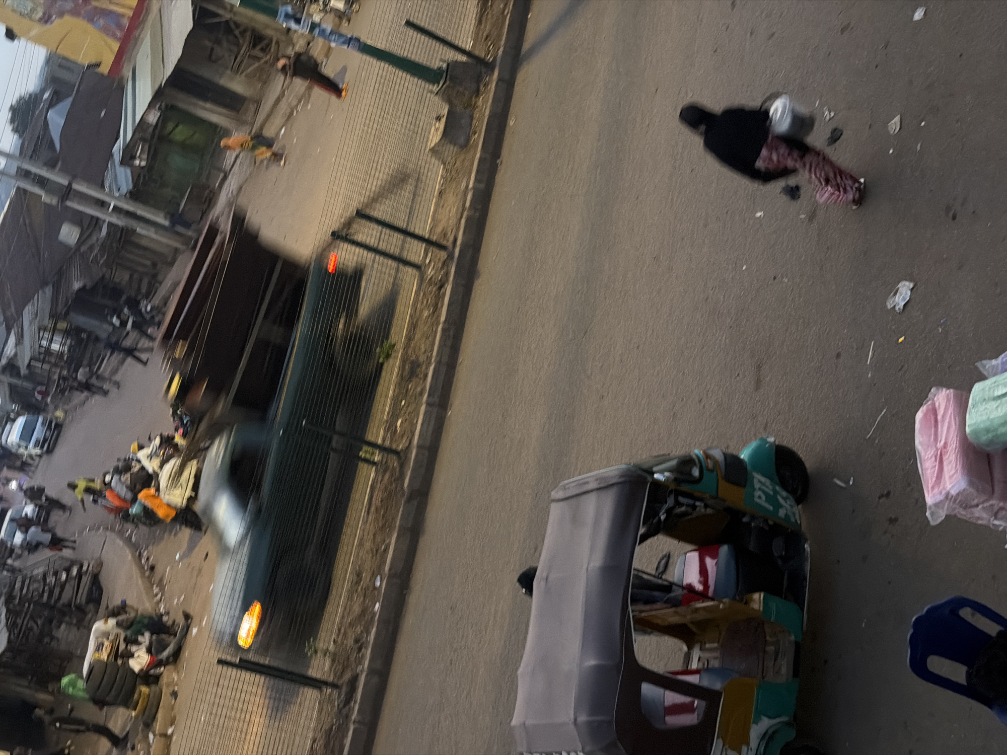 This image depicts a busy street scene in Bukuru, Nigeria. In the foreground, a colorful auto-rickshaw, identified by the "PTA" label, is parked on the asphalt road. A person, viewed from behind and slightly to the right, walks away from the rickshaw, carrying a metallic pail. Their attire consists of a black jacket and a patterned skirt.
Further down the road, a truck with a large, open cargo bed is visible, possibly engaged in waste disposal or transport. Several individuals are scattered around the street, some standing, others seated, and some appearing to be moving. A few buildings, appearing to be makeshift structures with corrugated metal roofs, line the street.
The overall impression is one of a typical urban environment in Nigeria, characterized by commercial activity and local transportation. The lighting suggests it is either early morning or late afternoon.