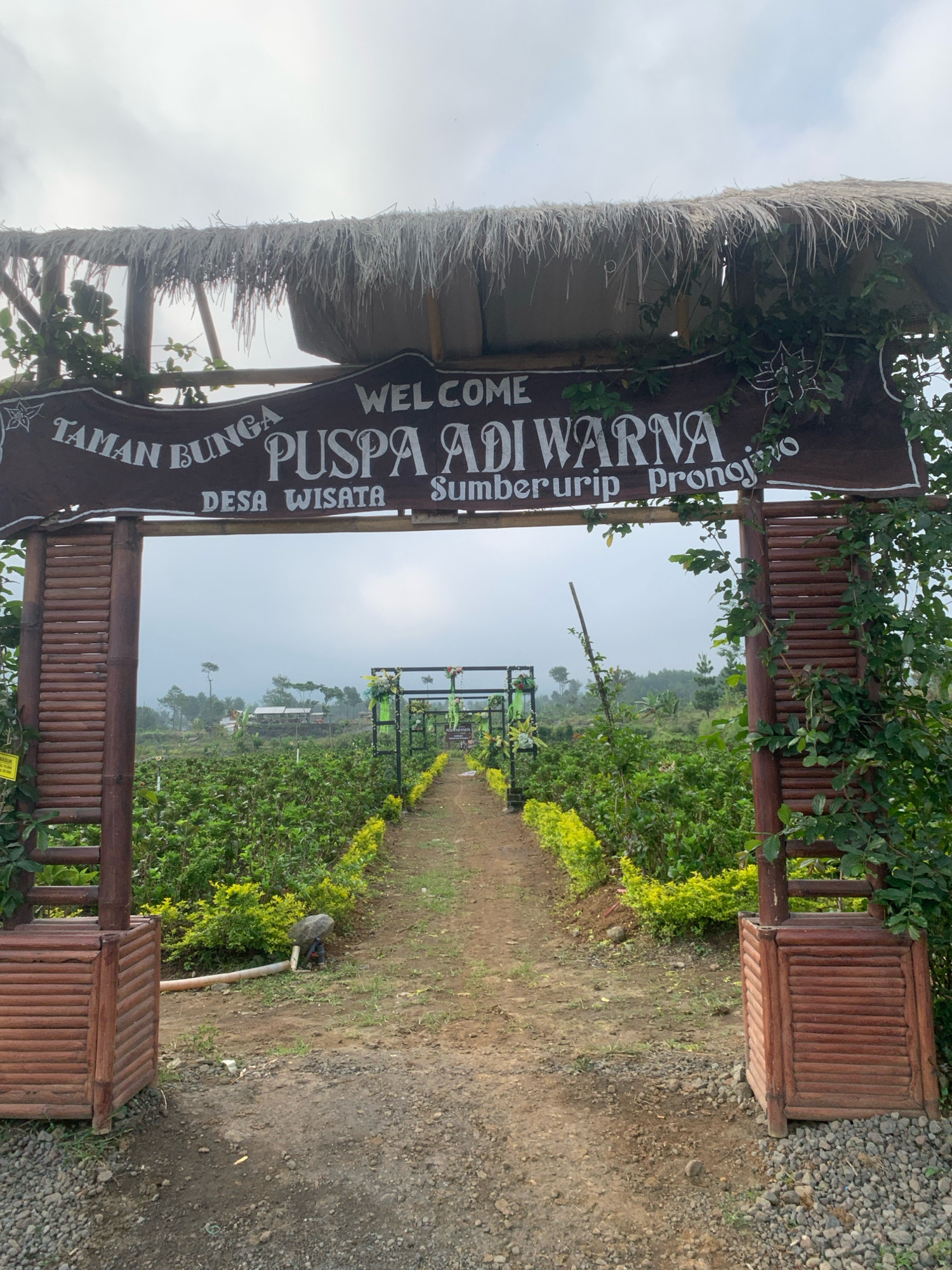 The image shows an entrance to a flower garden named "Taman Bunga Puspa Adi Warna" in Desa Wisata Sumberurip, Pronojiwo, Indonesia. A wooden archway with a thatched roof frames a dirt path leading into the garden. The path is lined with lush green plants and flowers. In the foreground, the entrance is decorated with bamboo elements and foliage. The sky is overcast.