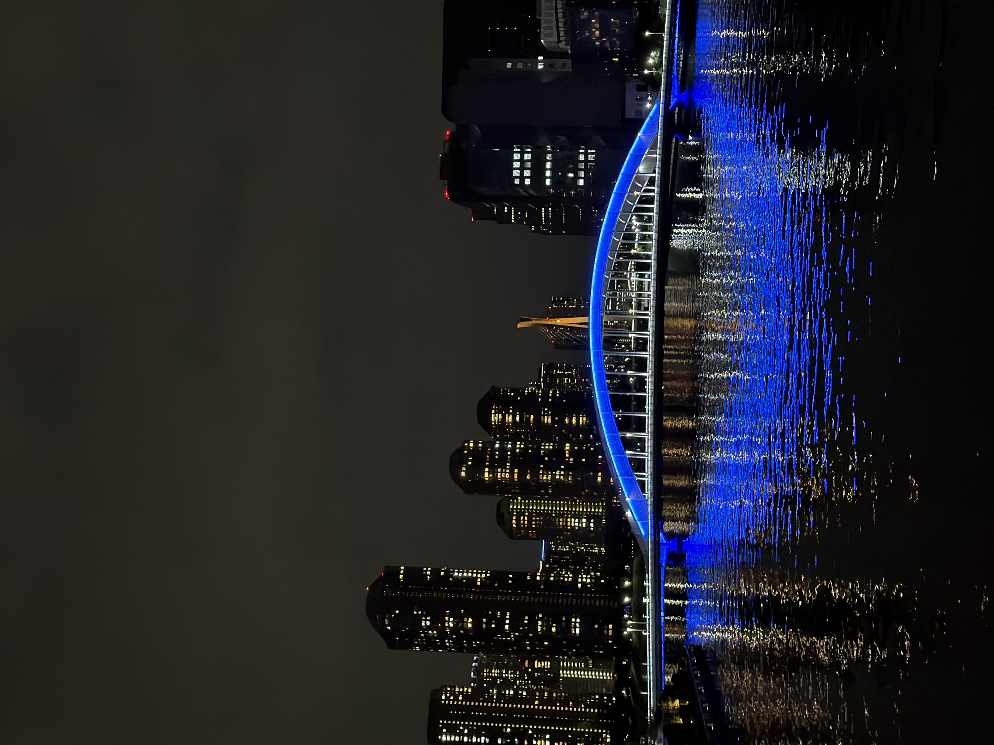The image shows a nighttime view of a modern city skyline with a brightly lit bridge over a body of water. The bridge is illuminated with vibrant blue lights, creating a striking reflection on the dark water. In the background, several tall buildings are visible, with numerous windows lit up, suggesting a densely populated urban area. The sky is dark and overcast, typical of nighttime in a city. The overall scene conveys a sense of urban dynamism and nighttime beauty. The location is identified as the Chuo City ward of Tokyo, Japan. Specifically, the image captures the Sumida River with the Kachidoki Bridge illuminated in blue.