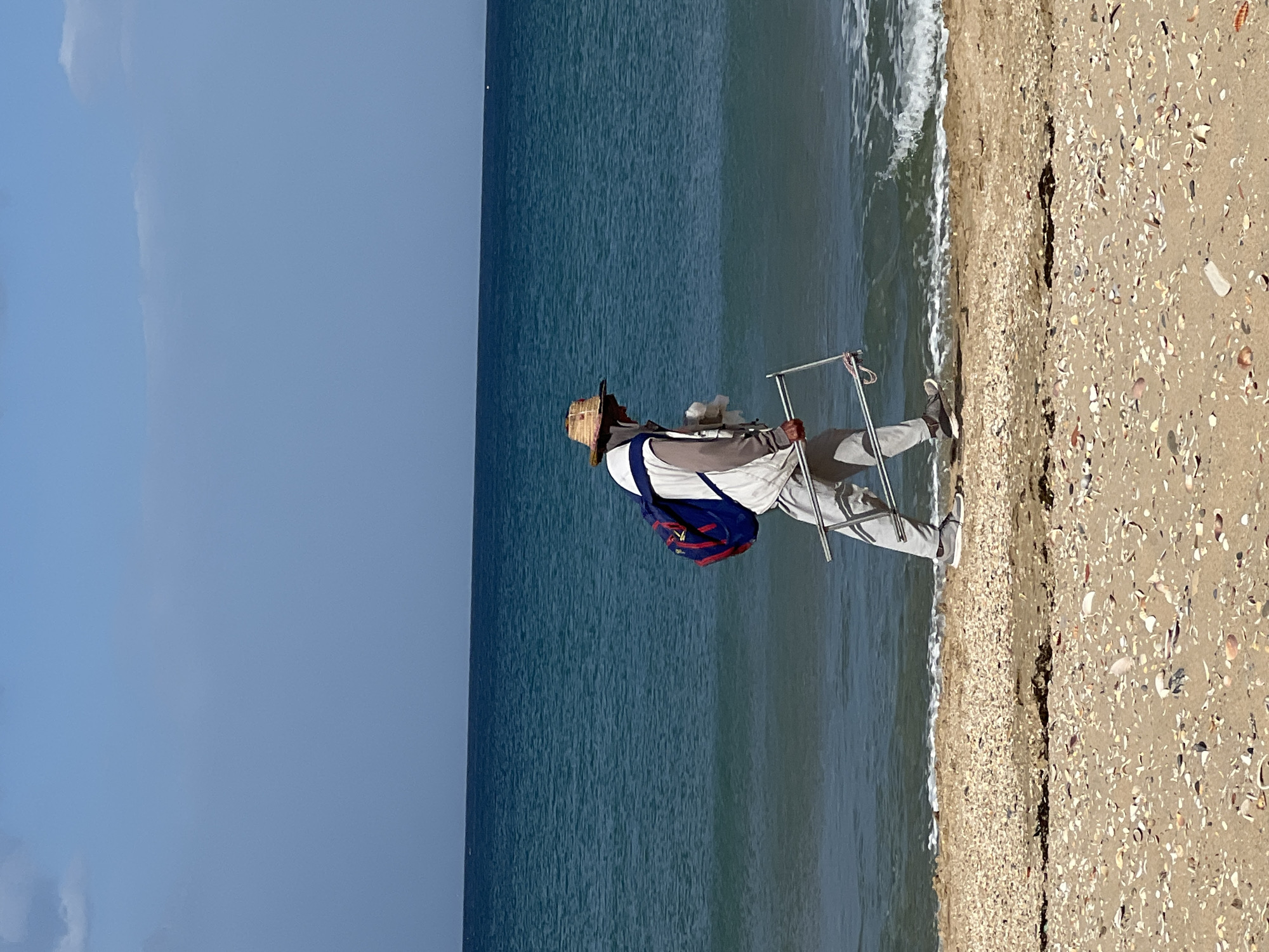 A man walks along a beach at M'Diq, Morocco. He wears a straw hat, light-colored pants, a white shirt with blue and gray accents, and carries a backpack and a metallic, folding object. The beach consists of sand and seashells. The ocean meets the shoreline with small waves. The sky above the ocean is clear blue.