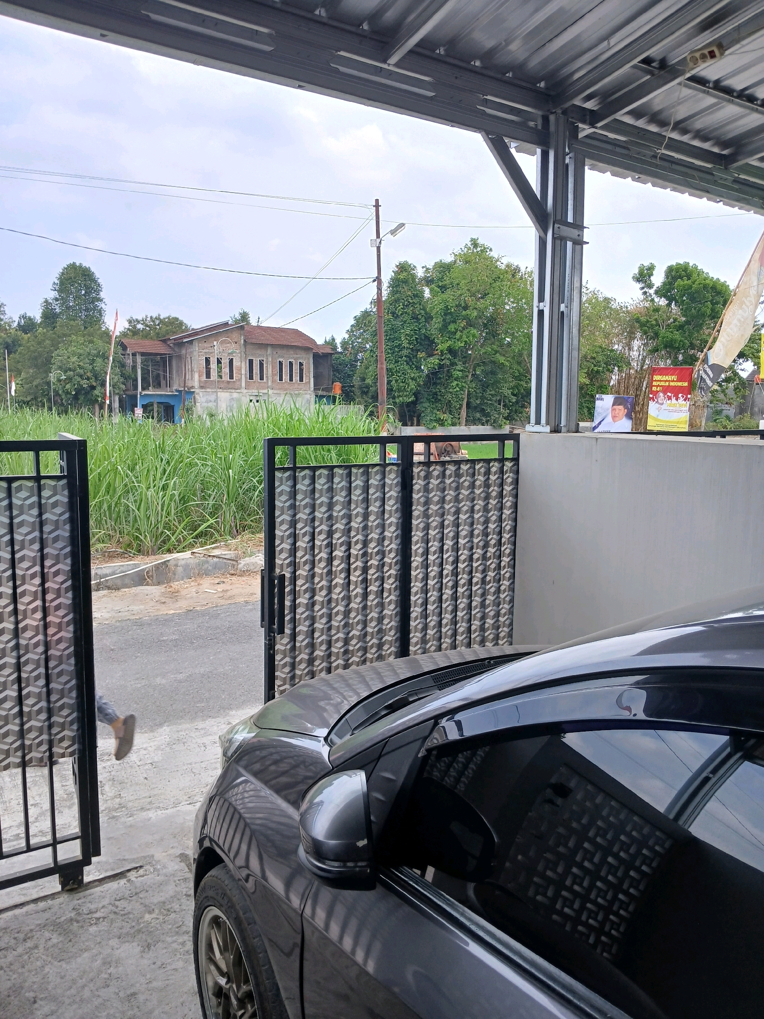 This image depicts an outdoor scene in front of a house located in Sitimulyo, Indonesia. A dark gray car is parked in the foreground, partially obscuring a grey concrete wall and a black metal gate with a patterned design. To the left of the gate, a person, whose legs and feet are visible, is walking away from the camera and towards the street. Beyond the gate, tall green grass is abundant, with a building under construction or renovation visible in the middle ground. The building has a reddish-brown roof and an unfinished facade with visible brickwork and arched window openings. A wooden pole with electrical wires runs across the scene, and a flagpole with an Indonesian flag is also present. In the background, trees and a cloudy sky complete the landscape. On the wall behind the gate, two posters are displayed; one is a red banner with white text, and the other is a smaller poster featuring a person's portrait. The overall environment appears to be a residential area with a mix of completed and unfinished structures.