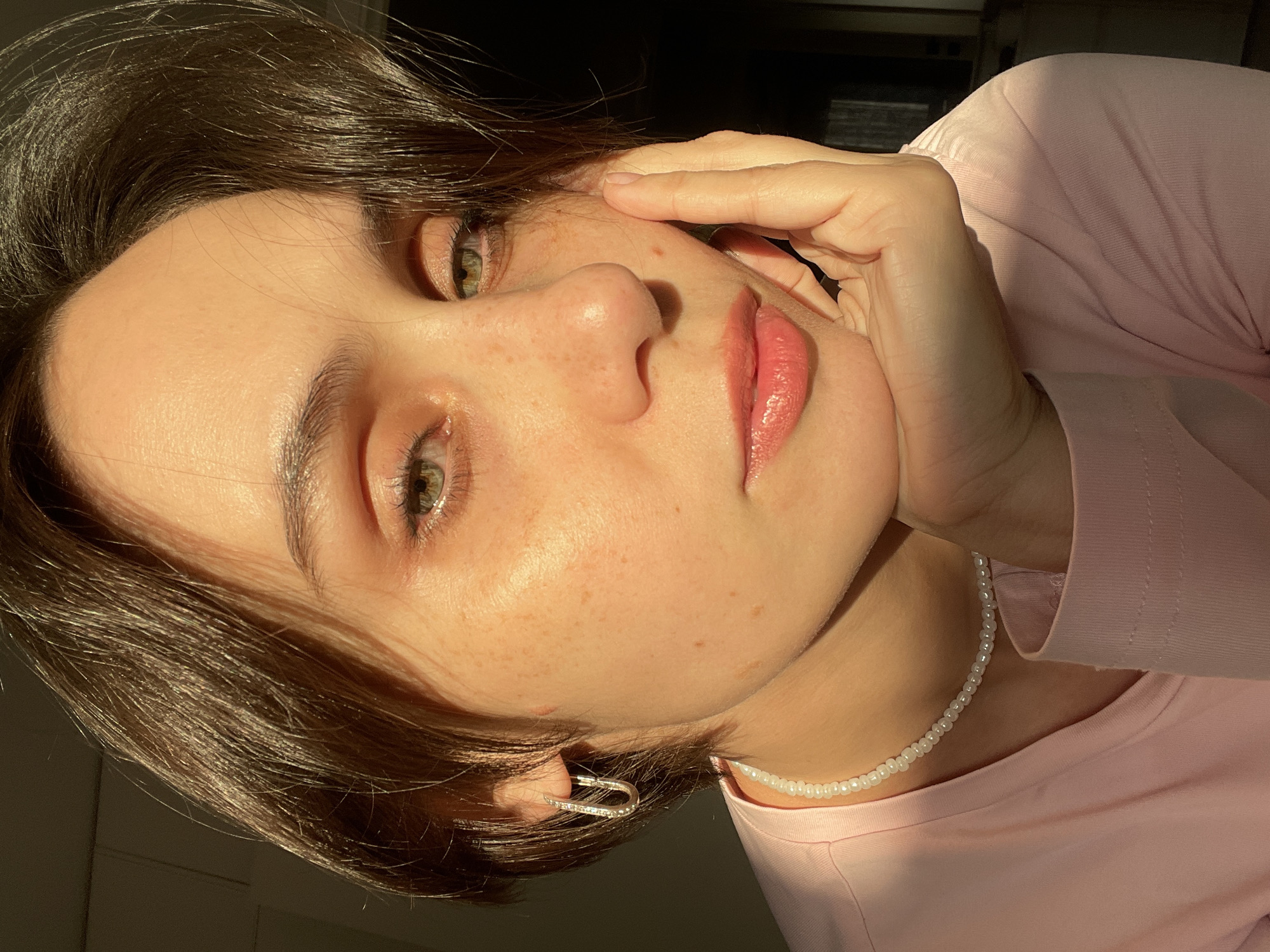 The image depicts a close-up, chest-up portrait of a single individual, appearing to be a young adult female, within an indoor setting. The specific location within Izhevsk, Russia, is not discernable from the provided visual information.
The individual has medium-dark brown, short hair, green eyes, and visible freckles across the face. They are wearing a light pink, long-sleeved top. A necklace composed of small, white, spherical beads is visible around the neck, and a silver-toned, U-shaped hoop earring, possibly studded with small clear stones, is worn in the visible left ear. The left hand is positioned with fingers gently resting against the chin and cheek. The individual is looking directly at the camera.
The scene is illuminated by strong, warm, directional light, likely natural sunlight, which highlights the right side of the subject's face (viewer's left) and hair, creating distinct shadows on the left side of the face and in the deep, indistinct background. The background is dark and out of focus, revealing minimal detail of the interior environment.