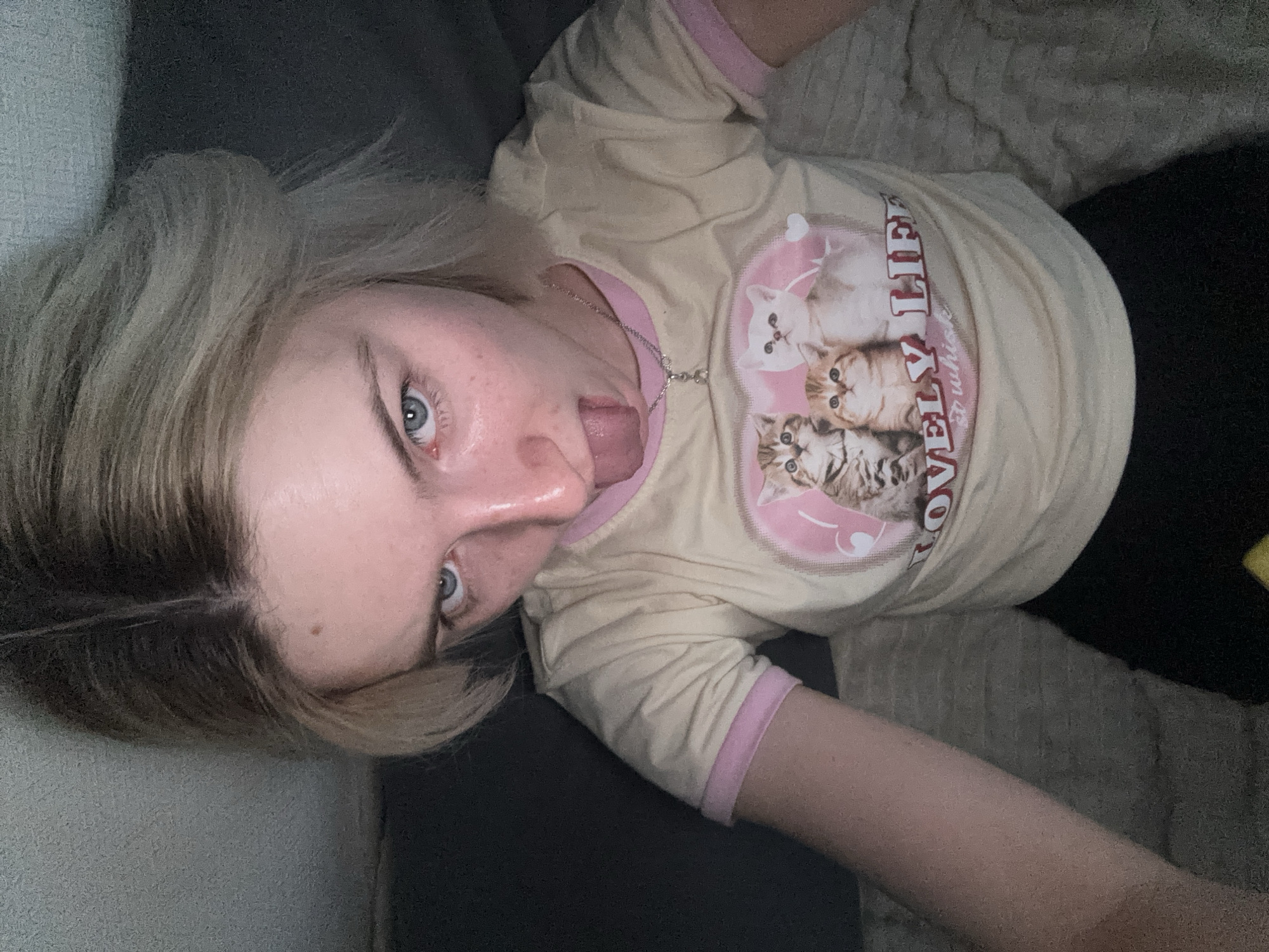 A fair-skinned individual with blonde hair is lying down, appearing to be on a sofa or bed. They are wearing a light yellow t-shirt with pink trim and a graphic featuring kittens. The individual's eyes are blue, and they are sticking out their tongue. A silver necklace is visible around their neck. The background consists of a dark gray sofa and a textured beige blanket. The location is provided as "Unknown, Russia."