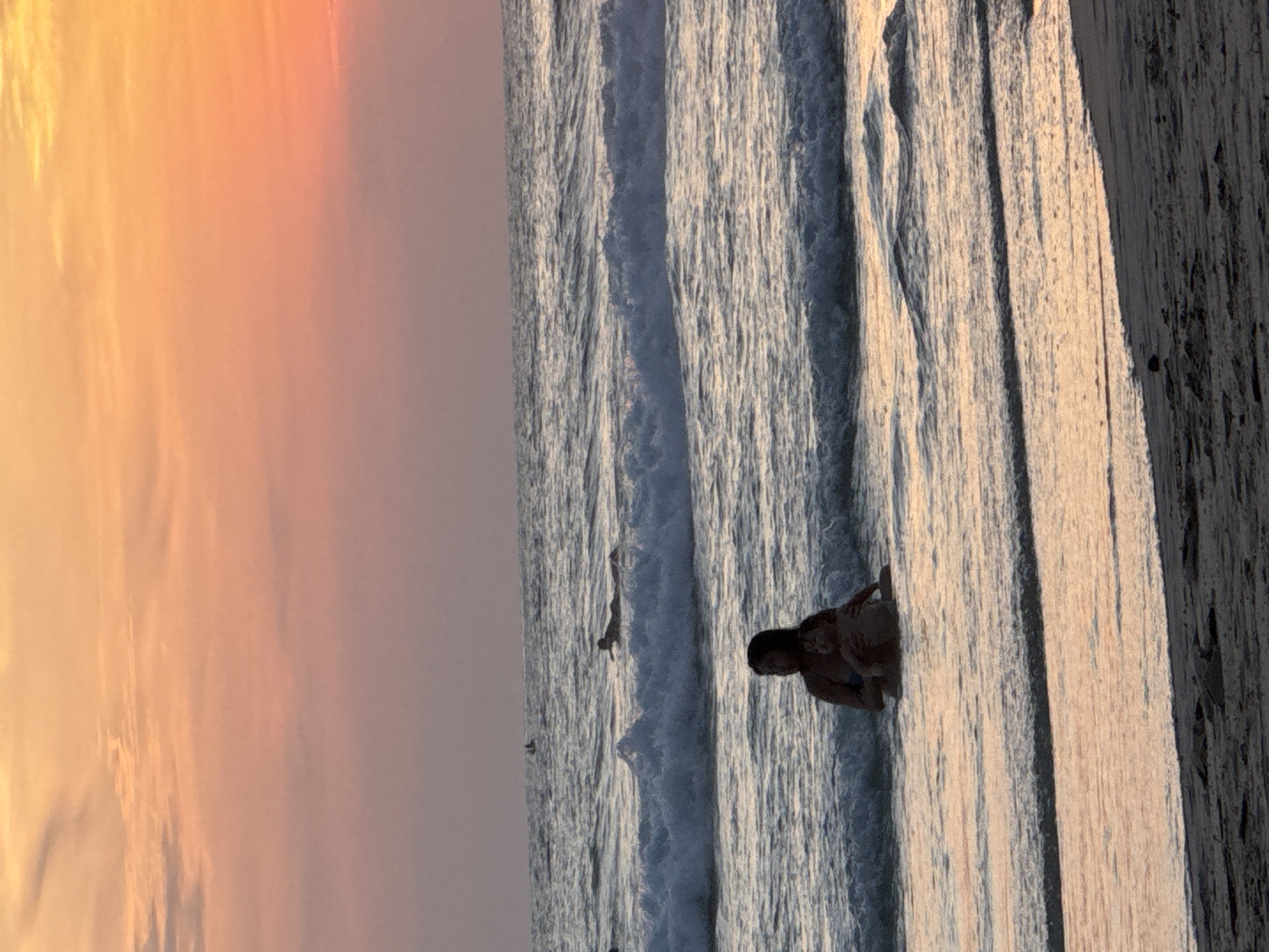A mother and child are sitting in the shallow water at Karon Beach, Thailand. The mother is wearing a blue bikini top and the child is being held in her arms. The sun is setting, casting a warm orange glow over the sky and water. Waves are gently rolling in towards the shore. In the distance, several surfers can be seen enjoying the waves. The sand is wet from the tide, and the overall atmosphere is peaceful and serene.