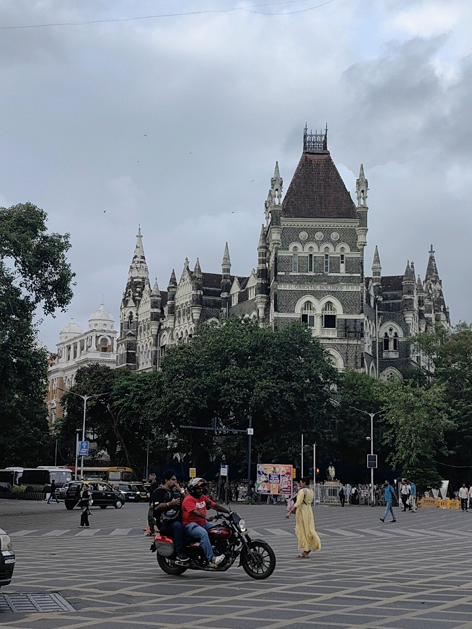 The image shows a view of the Oriental Buildings in Fort, Mumbai, India, on a cloudy day. The grand gothic-style architecture of the building is prominent, with its numerous spires, arched windows, and intricate stone carvings. In the foreground, a busy intersection with a pedestrian crossing is visible. Two people are riding a red motorcycle, with the rider wearing a red shirt and blue jeans, and the pillion rider in a black t-shirt and jeans, both wearing helmets. A woman in a long yellow dress walks across the pedestrian crossing. In the background, various vehicles, including buses and cars, are seen at the intersection. Trees line the streets, and streetlights are also visible. The sky is overcast with clouds.