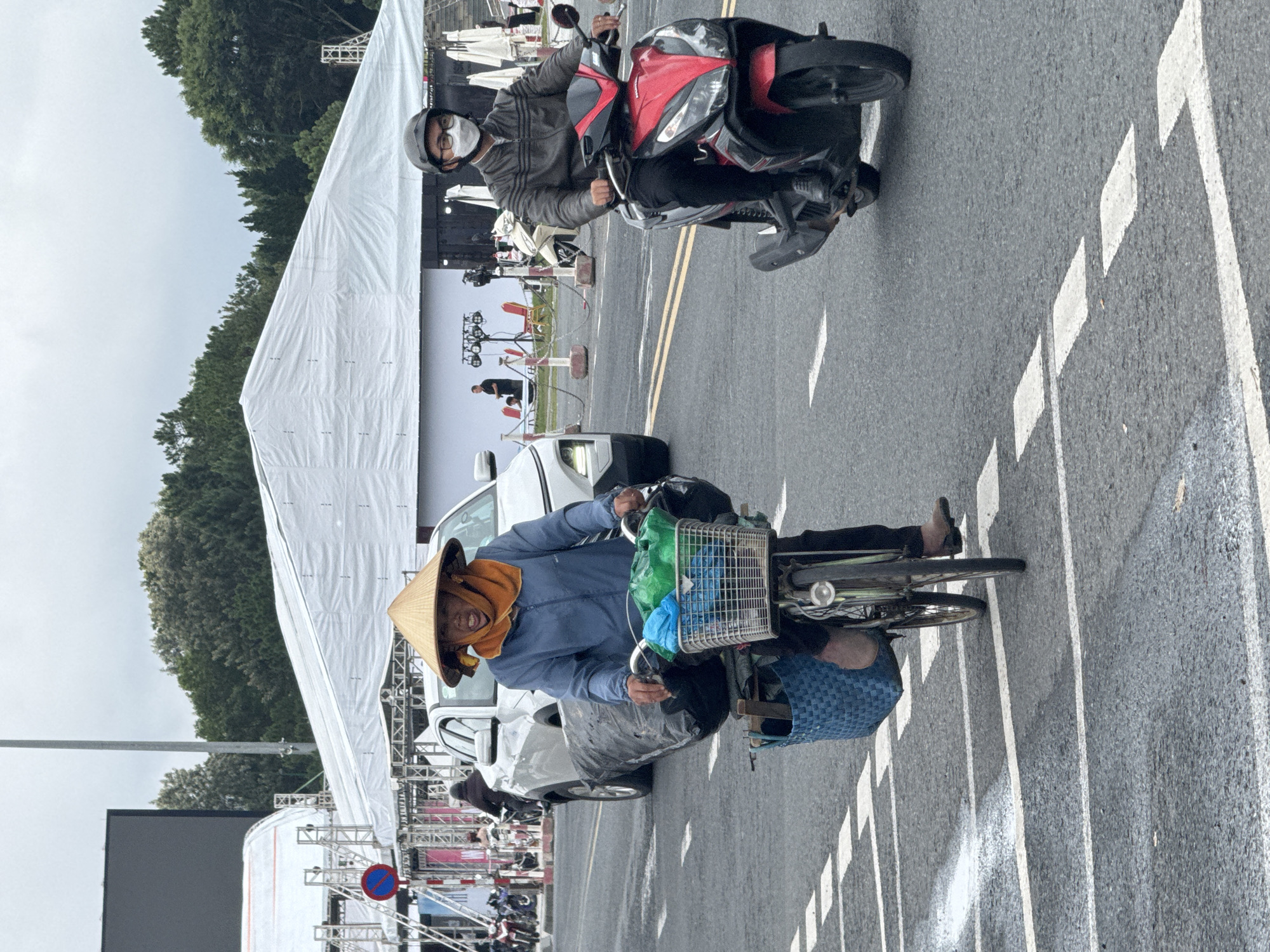 The image, oriented vertically, captures a moment on a road in what appears to be Da Lat, Vietnam, during an overcast day. Two individuals on two-wheeled vehicles are prominent, both looking towards the viewer.
In the foreground, slightly to the left, a person rides a bicycle. They wear a blue long-sleeved jacket and an orange scarf or towel around their neck, topped with a traditional conical hat (nón lá). A friendly smile is visible on their face. The bicycle is equipped with a wire basket at the front, holding green and blue items, and a blue woven bag hangs from the handlebars. The person's bare feet are visible near the pedals.
Behind the cyclist, and slightly to the right, another individual is on a red and black motor scooter. This person wears a grey jacket, dark trousers, a grey helmet, and a white face mask, obscuring part of their expression. They also appear to be looking towards the camera.
The road is asphalt, marked with white dashed and solid lines, including directional arrows/chevrons painted on the surface. In the background, large white temporary structures, possibly marquees or tents for an event, line both sides of the road. These structures are supported by visible metal scaffolding or trusses. Behind these, dense green trees are visible, extending towards an overcast sky. Parts of several white cars are also seen parked or moving behind the main subjects. A large, dark screen or display is visible in the distance, near the scaffolding on the left. A blue circular sign with a red diagonal line, indicating a prohibition, is discernible among the background structures. The scene suggests a public space, possibly set up for a specific event or festival.