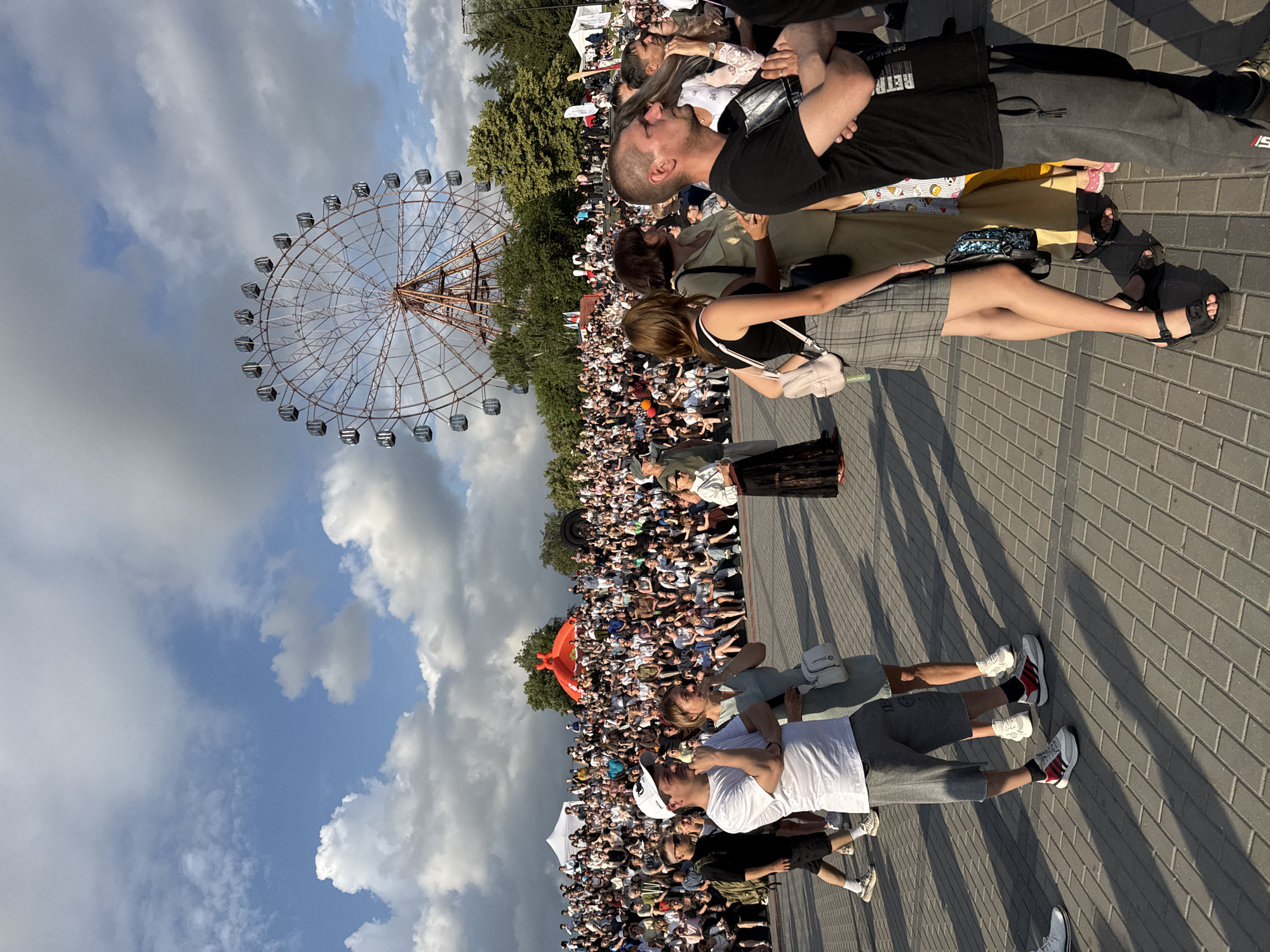 This image captures a large crowd gathered outdoors, likely for an event, with a Ferris wheel visible in the background against a cloudy sky. The scene appears to be a public space with paved ground. In the foreground, several individuals are visible, some looking in the same direction as if watching a performance or attraction. The crowd is densely packed, occupying a large area and seated on tiered risers in the background. The overall atmosphere suggests a daytime event in a city park or fairground. This scene is consistent with an event taking place at the Novosibirsk Central Park.
