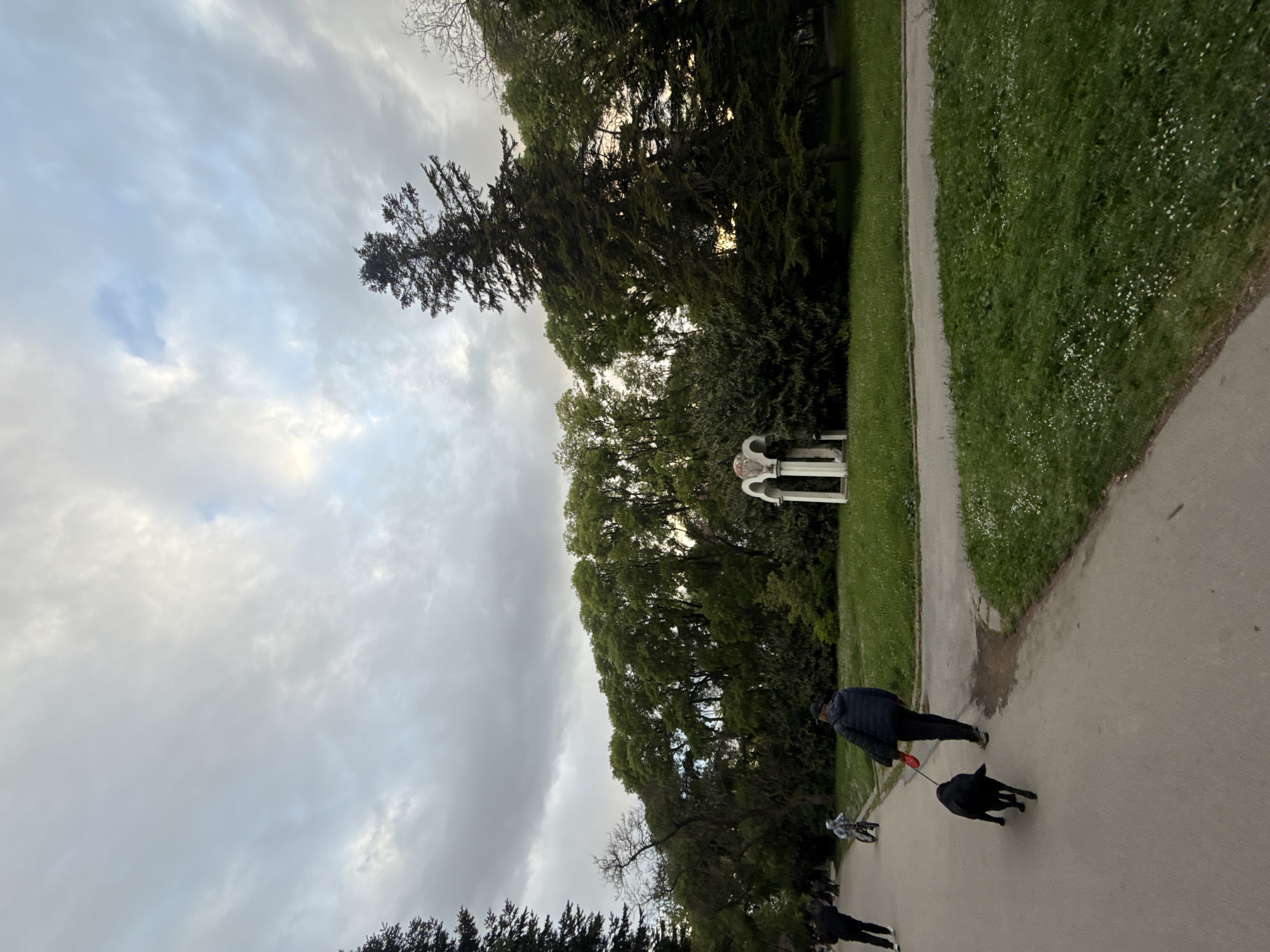 A park scene under a cloudy sky shows a paved path alongside grassy areas and trees. A person wearing a dark jacket walks a black dog on a red leash. Further down the path, another person is seen. A white architectural structure is visible amidst the trees. The location is not explicitly stated as Varna, Bulgaria; it is a park setting based on the provided image.