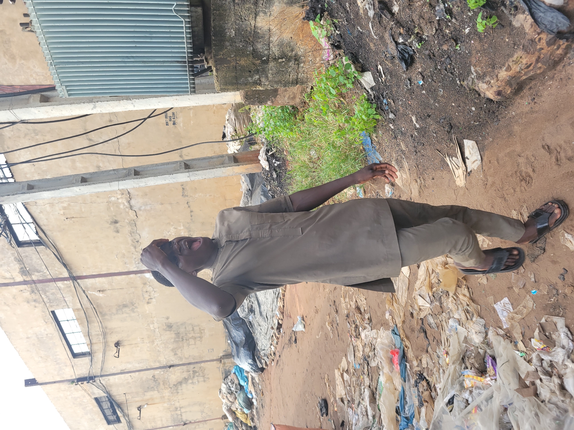 An image shows a man outdoors in what appears to be an urban environment, potentially in Onitsha, Nigeria, based on the prevalence of open garbage and the style of clothing. He is wearing a long-sleeved shirt and trousers. The man appears to be looking up, with his mouth open and hand to his head. The background includes buildings with visible windows and electrical wires, as well as significant amounts of discarded plastic and other types of waste. The ground is dirt and littered with debris.