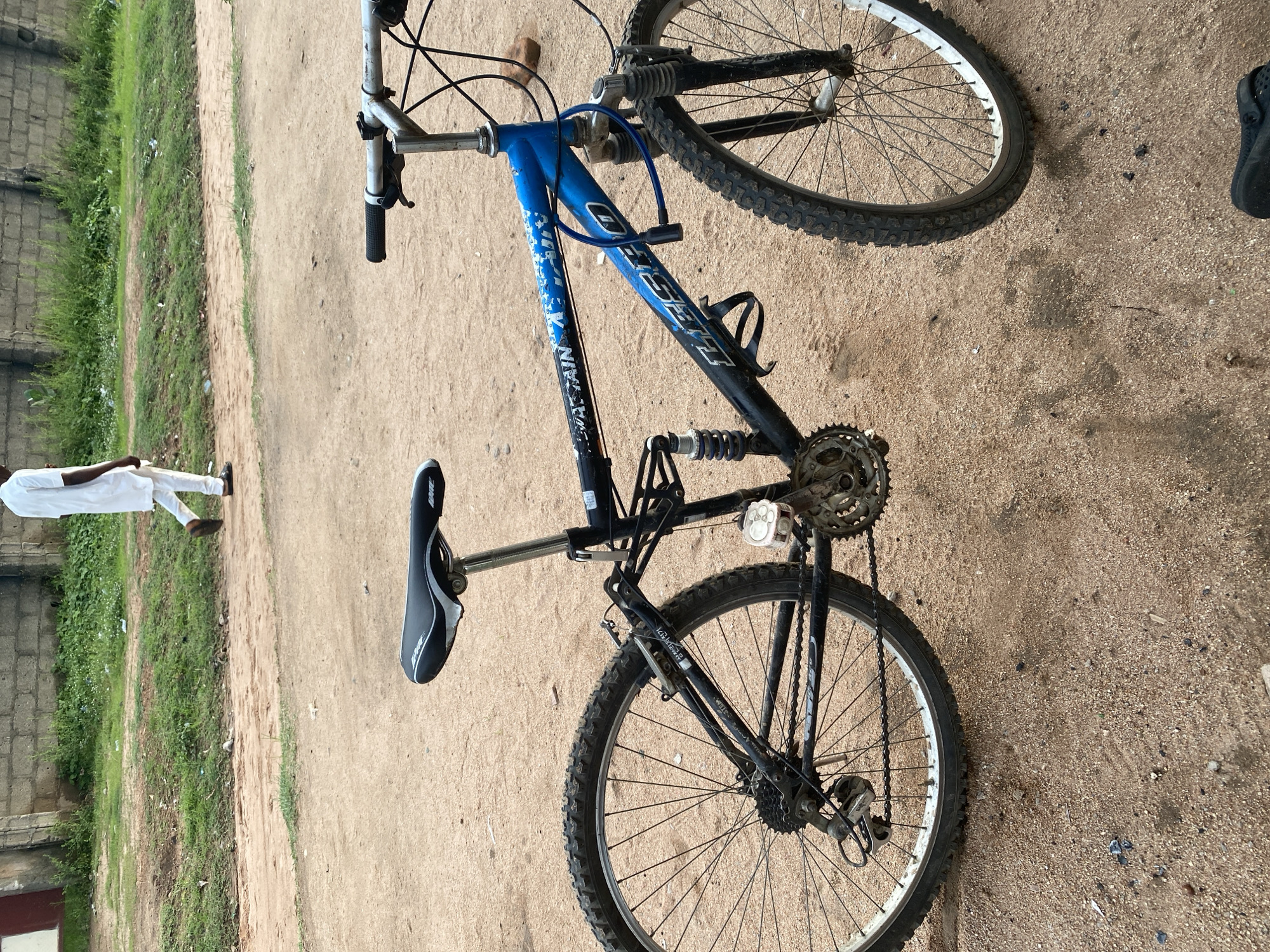 A full-shot, eye-level photograph depicts a blue and black full-suspension mountain bicycle parked on a dirt path in Jalingo, Nigeria. The bicycle is positioned diagonally, with its front wheel to the right and rear wheel to the left. The frame is primarily black with blue accents and white lettering that reads "MOUNTAIN BIKE" and "LIBRO." The handlebars are silver with black grips. A blue cable lock is attached to the frame. The rear derailleur and chain are visible. The seat is black with white trim.
In the background, a person in a white tunic and trousers walks away from the camera on the same dirt path, which borders a grassy area. Beyond the grass, a low, rough brick wall with some vegetation growing on top is visible. The environment appears to be an outdoor, somewhat undeveloped urban or semi-urban area. The lighting suggests it is daytime, possibly late afternoon or early morning, with soft shadows.