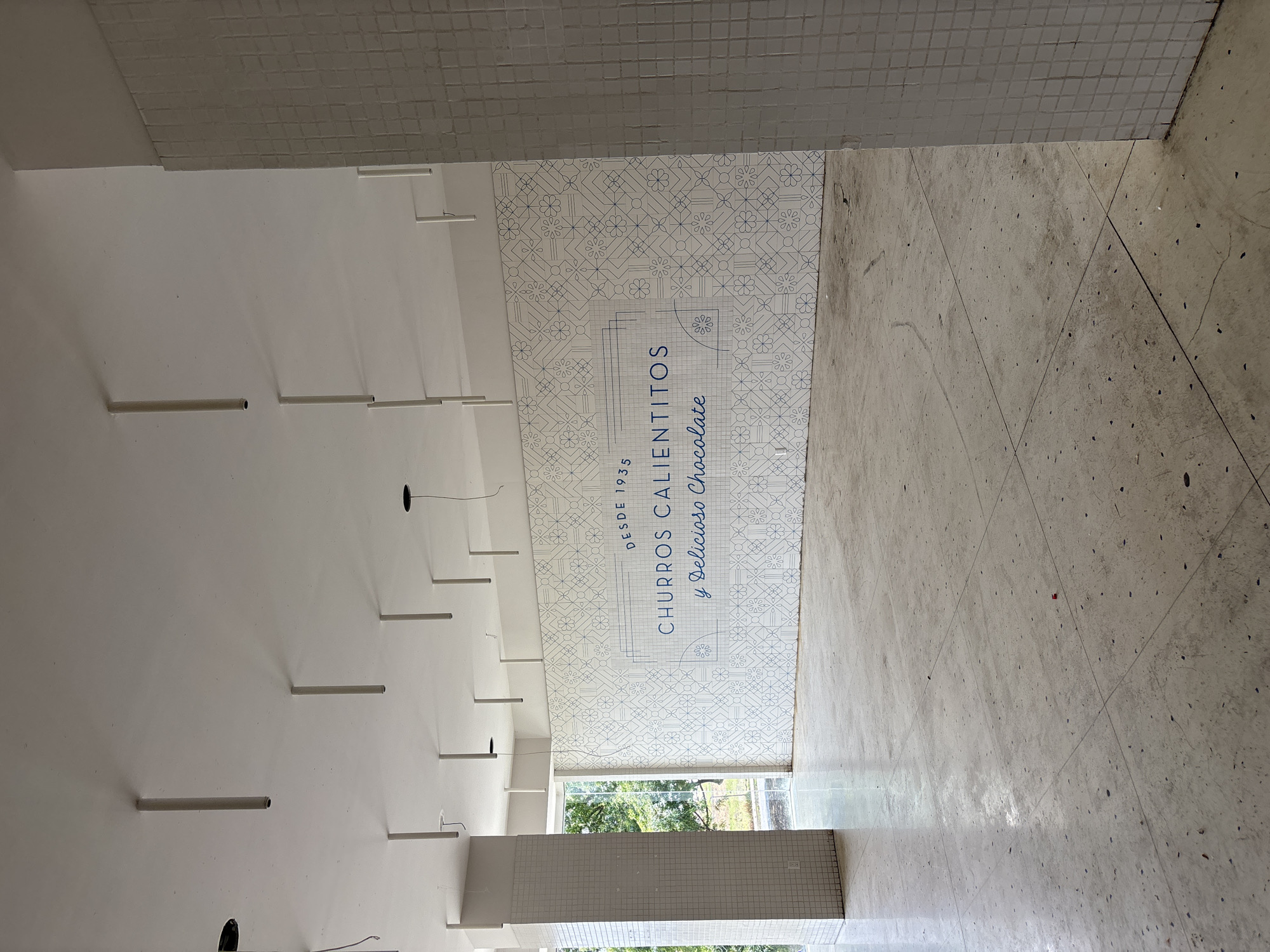 The image shows the interior of a building in Metepec, Mexico. The walls are primarily white, with a decorative panel featuring a geometric design and the words "CHURROS CALIENTITOS y Delicioso Chocolate DESDE 1935". The floor is tiled with large, light-colored tiles. The ceiling has a series of cylindrical and rectangular elements. A window or doorway is visible at the far end of the space, revealing greenery outside. There are no people visible in the scene.