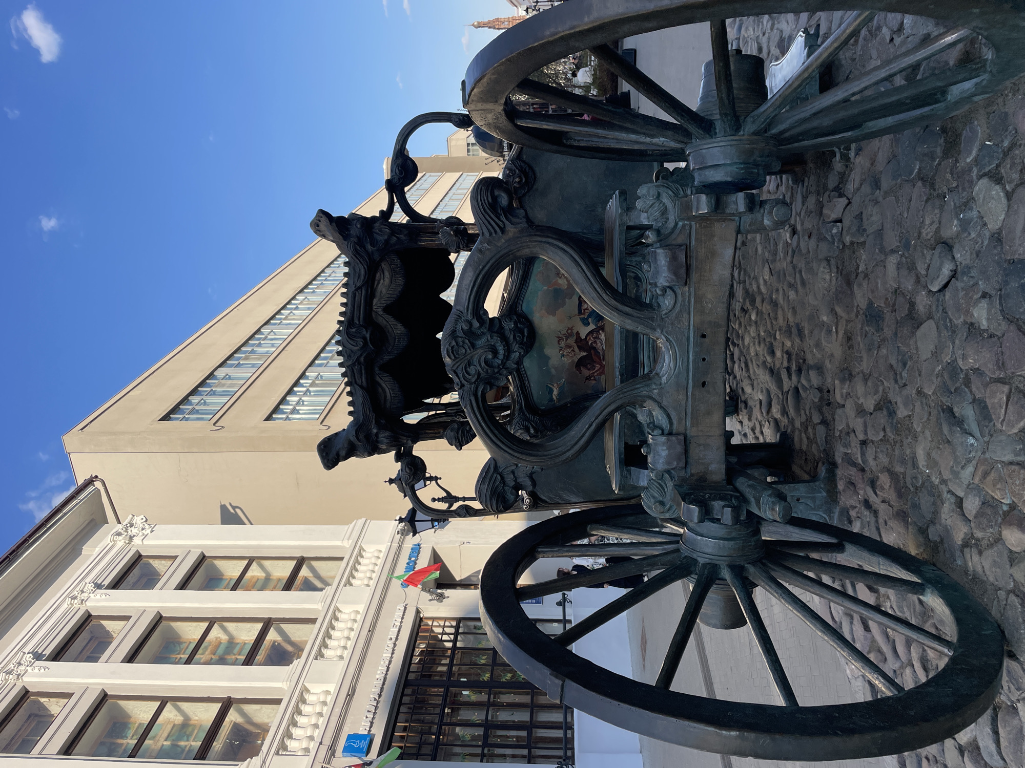 A low-angle shot depicts a bronze carriage on a cobblestone street in Kazan, Russia. The carriage is elaborately designed with ornate scrollwork and animalistic sculptures. It appears to be a public art piece, likely a monument. The frame is dominated by the carriage's large wheels, which are made of metal. The building facades on either side of the carriage are modern with rectangular windows. A clear blue sky is visible in the background. A small building has colorful flags displayed outside. The cobblestone pathway on which the carriage rests is uneven and textured.
