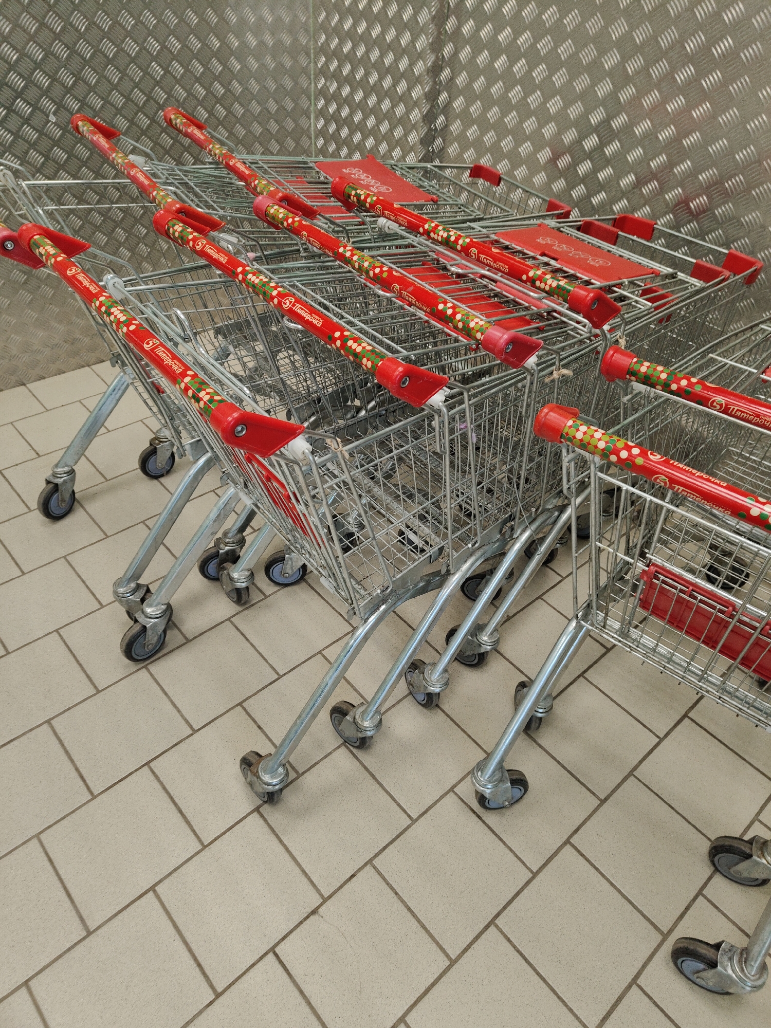 The image shows a row of shopping carts with red handles and the logo "5 Пятерочка" (5 Pyaterochka) stacked together in what appears to be a storage area. The location is unknown. The carts are made of metal wire. The background is a metal wall with a diamond pattern. The floor is tiled in light beige. The carts are empty and not in use.