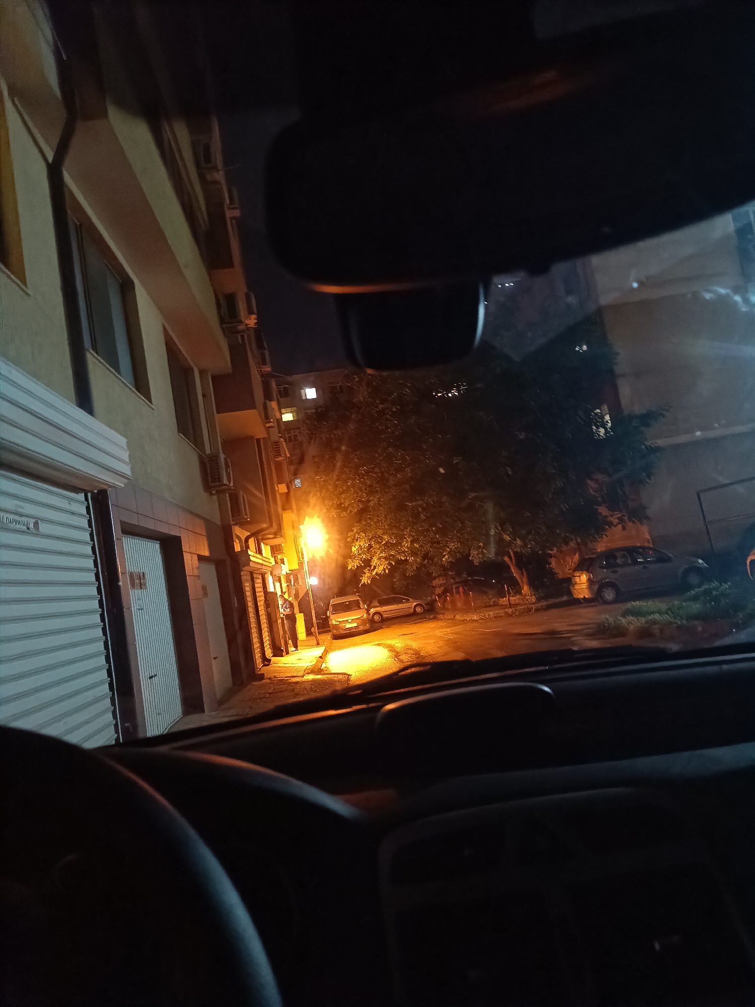 The media file captures a nighttime street scene from the perspective of someone inside a vehicle, looking out through the windshield. Parts of the car's dashboard, a segment of the steering wheel on the lower left, and the rearview mirror at the top are visible in the foreground.
The scene outside features a narrow, dimly lit street lined with multi-story buildings and trees. On the left, a light-colored building displays closed roll-down shutters on its ground floor, likely for garages or commercial spaces. Several air conditioning units are mounted on the facades of the buildings. Windows in some upper-floor apartments are illuminated, indicating presence.
A bright, warm-toned streetlamp on the left side of the street casts a strong glow on the immediate vicinity. A male figure is visible standing near this streetlamp, partially illuminated and dressed in dark clothing. Further down the street, several parked cars are discernible, including a light-colored hatchback on the right. Dark, dense foliage from trees adds to the urban landscape. The sky above is dark, consistent with nighttime.
The atmosphere is quiet and urban, suggesting a residential or mixed-use area in Varna, Bulgaria. On a white roll-down shutter on the far left, there is faint Cyrillic text, but it is largely illegible due to the image resolution and distance.
