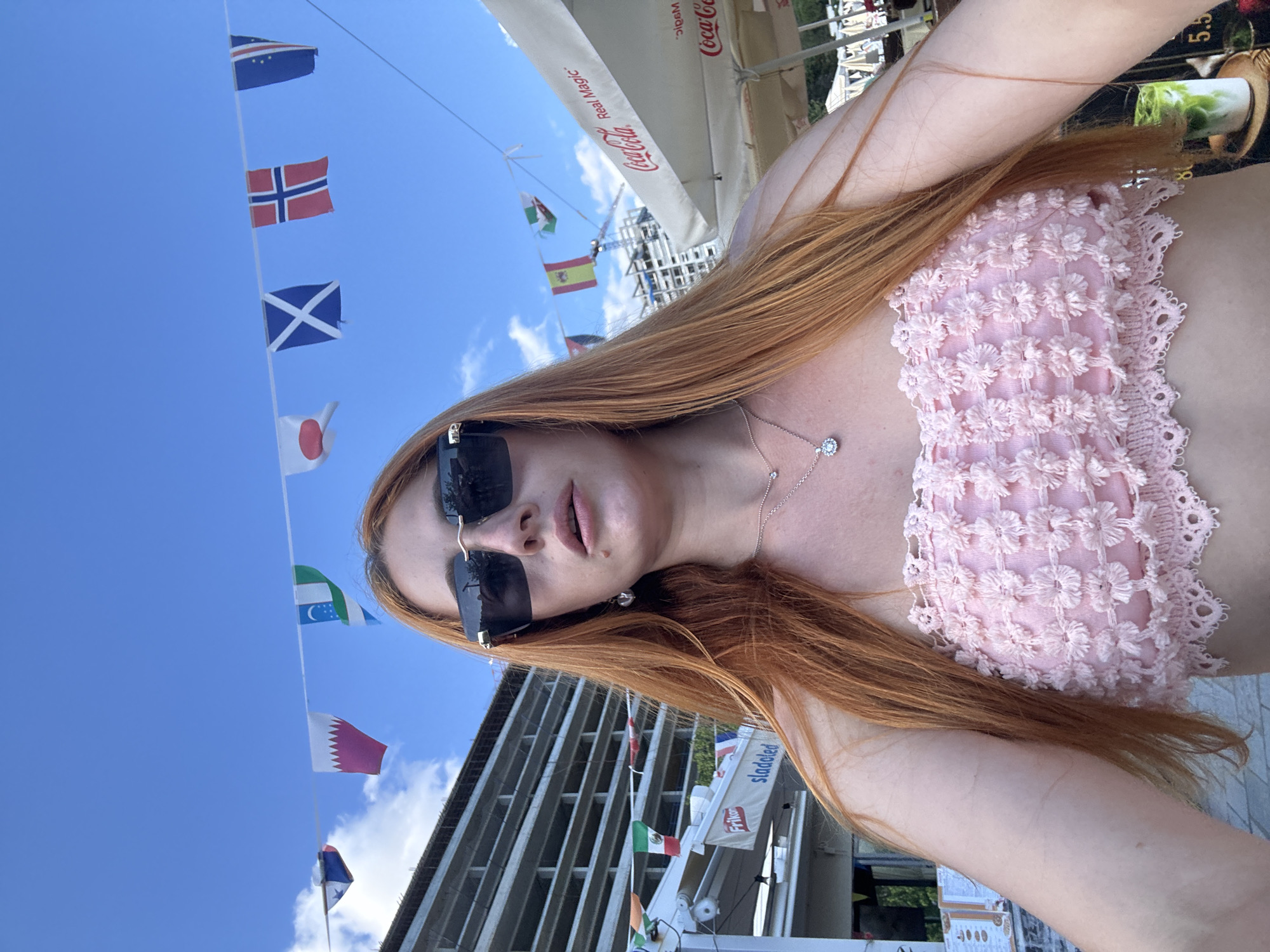 The image captures a selfie of a woman with long, reddish-brown hair and dark sunglasses, taken outdoors on a bright, sunny day. She is wearing a light pink, intricately detailed crop top with a lacy or embroidered texture. A delicate silver necklace with a small pendant adorns her neck.
The background features a clear blue sky crisscrossed by lines from which numerous national flags are strung. Identifiable flags include those of Cape Verde, Norway, Scotland, Japan, Spain, Wales (partially visible), Qatar, Mexico, and Ireland, suggesting an international or maritime theme. White outdoor umbrellas, one branded with "Coca-Cola" and "Real Magic," indicate an outdoor cafe or restaurant setting. A modern building facade and other structures are visible in the distance, along with a partial view of a sign displaying "Sladolèd" (ice cream) and a Frikon logo. Part of a menu or price list showing "5.5" is also visible in the upper right corner.
The scene depicts a leisurely outdoor moment, likely at a vibrant tourist location like Budva, Montenegro, given the array of international flags and cafe setting. The bright daylight suggests it is daytime.