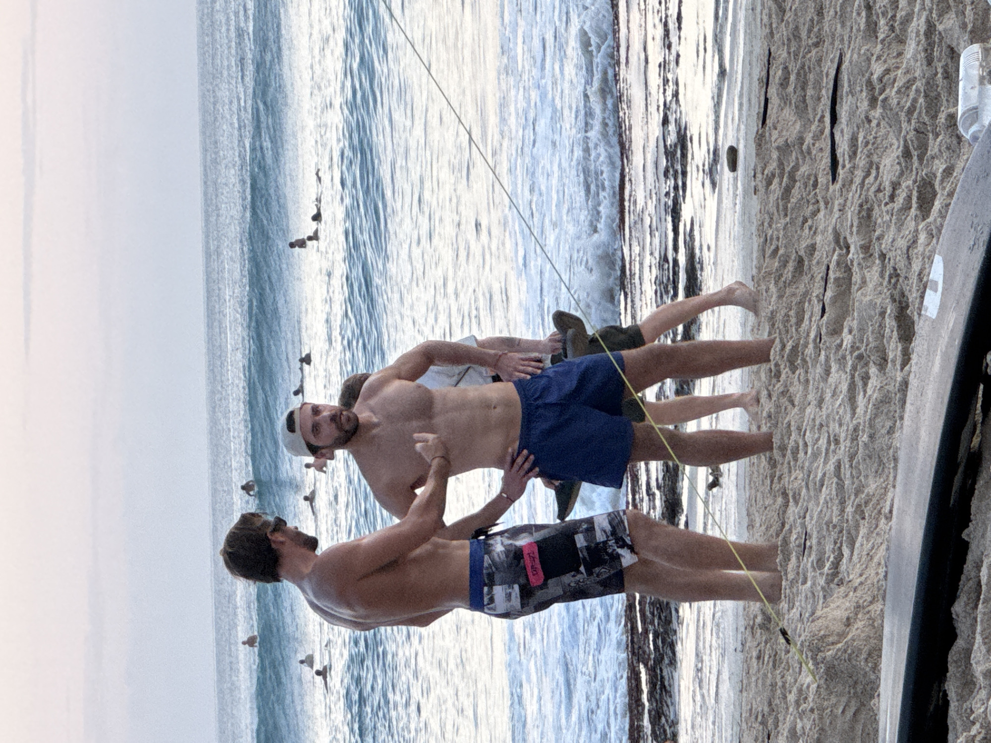 At the beach in Kuta Selatan, Indonesia, two men stand on the sand near the water's edge. The man on the left wears patterned swim trunks, while the man on the right wears blue swim trunks and a baseball cap. Both are shirtless. The ocean is visible behind them, with several surfers in the water. The sky is overcast. A surfboard lies in the foreground on the sand.