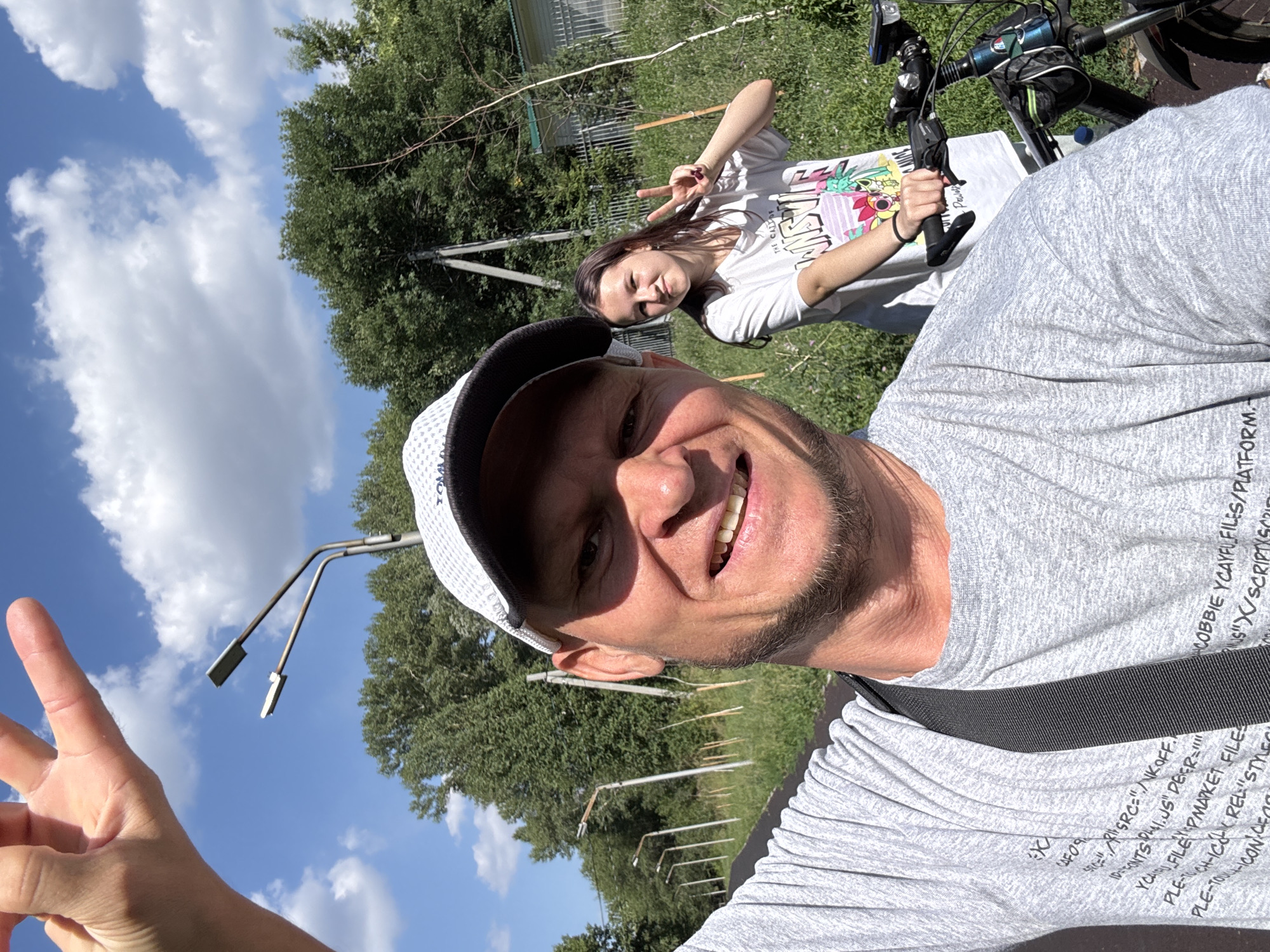 In this selfie, a man in his 30s or 40s, wearing a grey t-shirt and a white cap, smiles broadly at the camera. His right hand is raised, with fingers extended, as if waving. Behind him, a teenage girl, with long brown hair, also smiles and makes a peace sign with her right hand. She is wearing a white t-shirt with a colorful graphic design. They are standing next to a bicycle.
The background suggests an outdoor setting, possibly a park or a recreational area, with a paved path in the foreground. Trees and green grass are visible, and a few streetlights line the path. In the distance, there is a fence and a building. The sky is blue with scattered white clouds, indicating a sunny day.
The image appears to have been taken in Oskemen, Kazakhstan, based on contextual information provided. Specifically, the presence of streetlights and the natural environment are consistent with a park or a suburban area within the city.