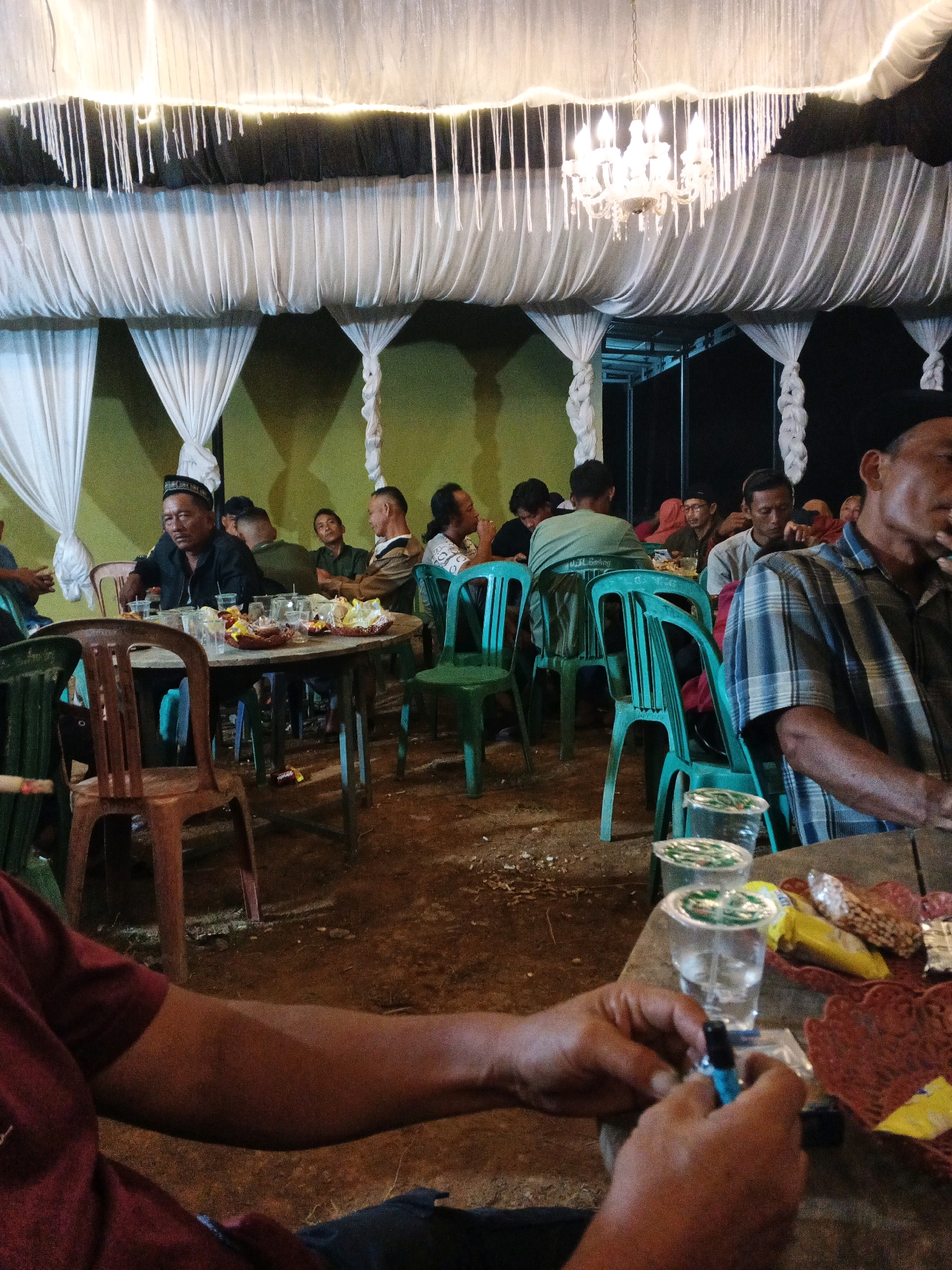 The image captures an indoor gathering, likely a social event, taking place in Brangan Mulya, Indonesia. The scene is dimly lit, with a festive ambiance created by white drapes hanging from the ceiling, adorned with small lights and a chandelier. The ground appears to be dirt, suggesting an outdoor or semi-outdoor setting.
Several individuals are seated around round tables, engaged in conversation and eating. Many are men, some wearing traditional Indonesian attire like peci hats. The tables are laden with food items, snacks in baskets, and drinks in plastic cups. The presence of plastic chairs indicates a casual setup. In the foreground, a person's arm and hands are visible, holding a small object, possibly a lighter, and a cigarette. The overall impression is of a community event, perhaps a celebration or a religious gathering, characterized by a communal dining experience and social interaction. The background reveals more seating and people, further emphasizing the communal nature of the event.