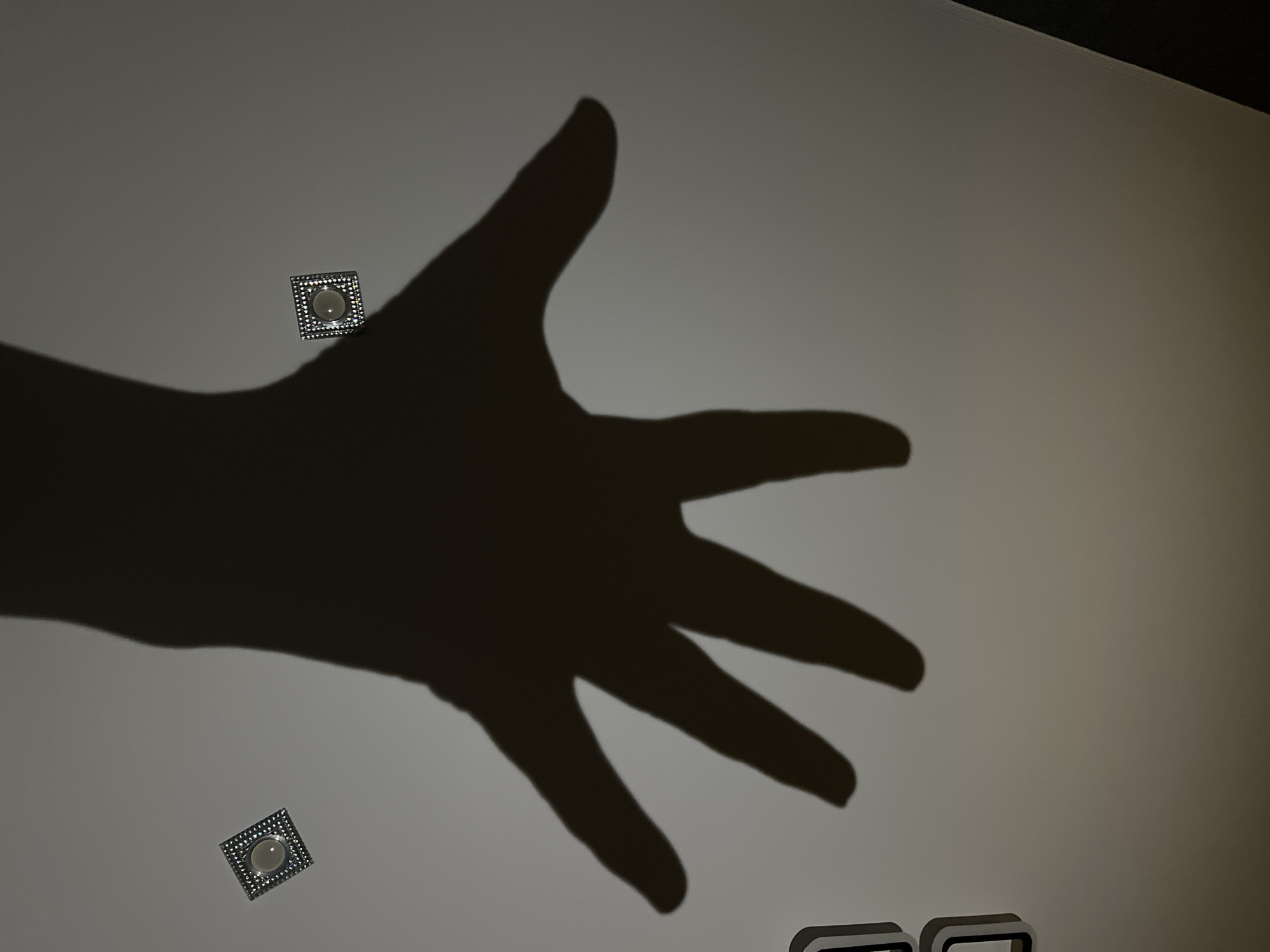 The image shows the shadow of a hand projected onto a light-colored wall. Two square light fixtures are visible on the wall, and the bottom of two rectangle fixtures as well. There is no information available to establish a location in the city of Chaikovsky, Russia.