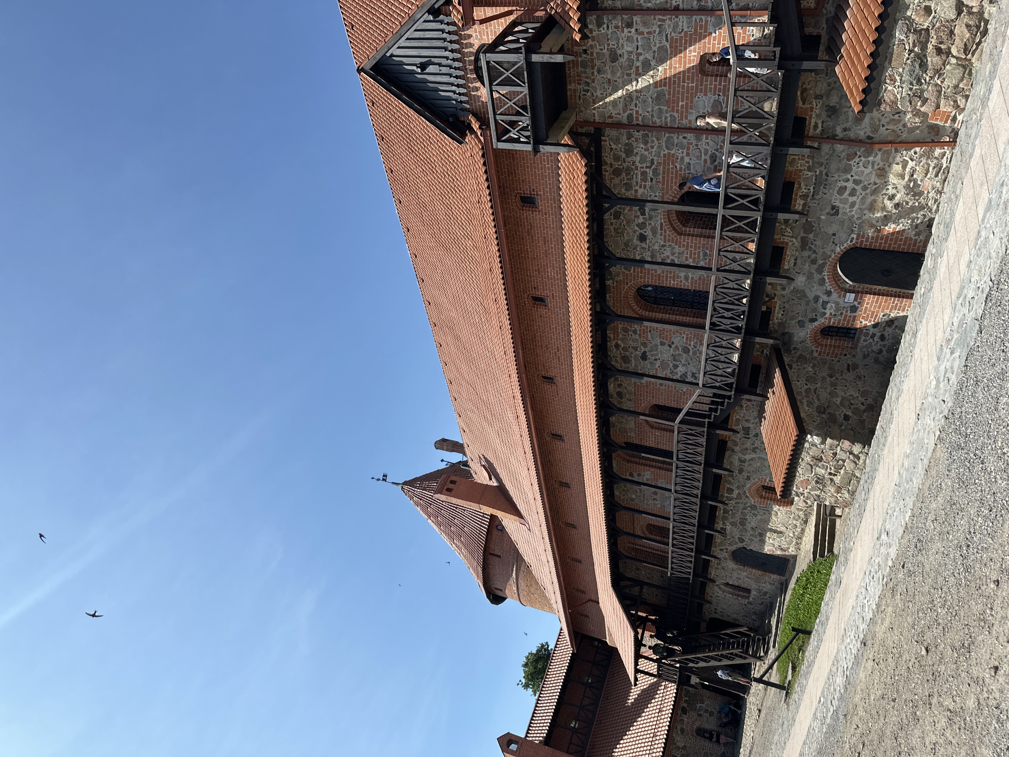 The image depicts the Trakai Island Castle in Trakai, Lithuania. The castle is constructed primarily of red brick and stone, with distinctive terracotta tiled roofs. A wooden walkway with intricate railing details runs along the upper levels of the castle, connecting various sections. Several people are visible on the walkway, some walking and others conversing. In the background, a conical tower with a flag at its peak rises above the main structures. The sky above is a clear, bright blue with two birds in flight. The ground in the foreground is a mix of gravel and paved pathways. The overall scene suggests a sunny day, likely during tourist season, with visitors exploring the historical landmark.