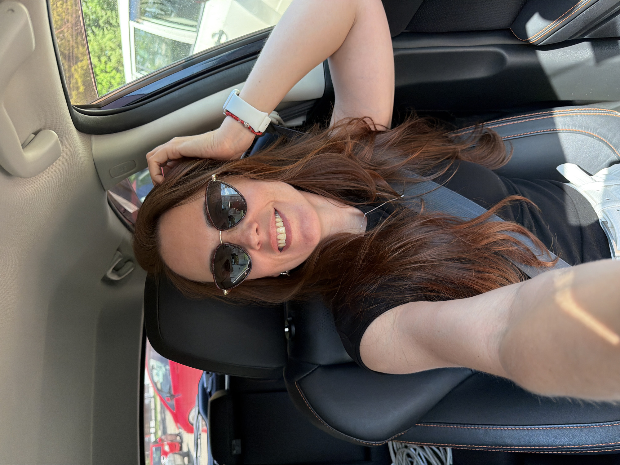 A woman with long, auburn hair is taking a selfie in the driver's seat of a car. She is wearing sunglasses and a black shirt. Her right arm is raised, with her hand resting on her head. She is also wearing a white smartwatch and a red beaded bracelet on her wrist. The interior of the car is visible, including black leather seats with orange stitching. Outside the car, other vehicles and greenery can be seen. The location is Birobidzhan, Russia.