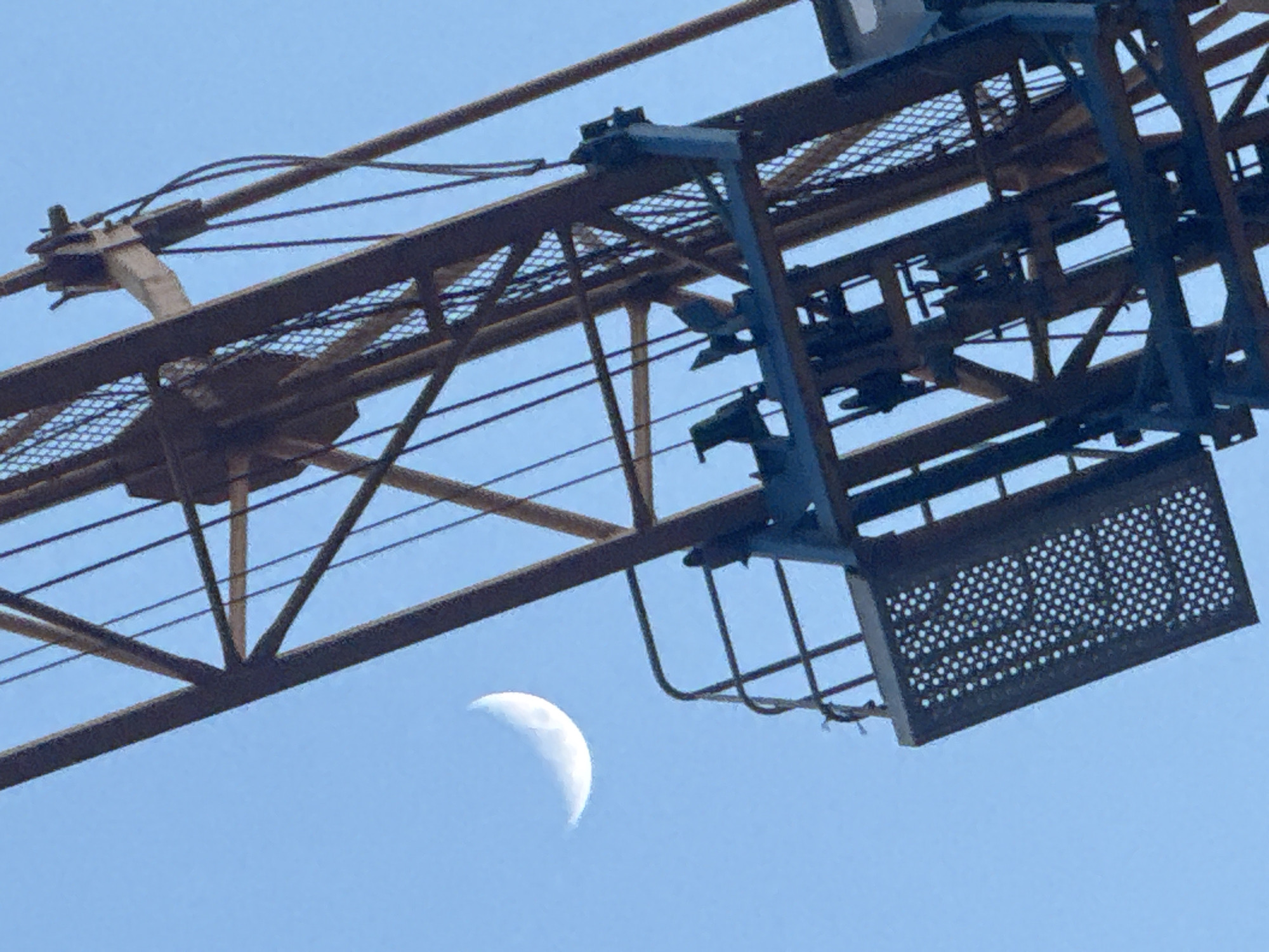 Captured from Ramat Gan, Israel, the image features a partial view of a crane juxtaposed against a pale blue sky. The crane's structure dominates the foreground, revealing its intricate metal framework. A waning crescent moon is visible in the lower left, positioned near the crane's base.