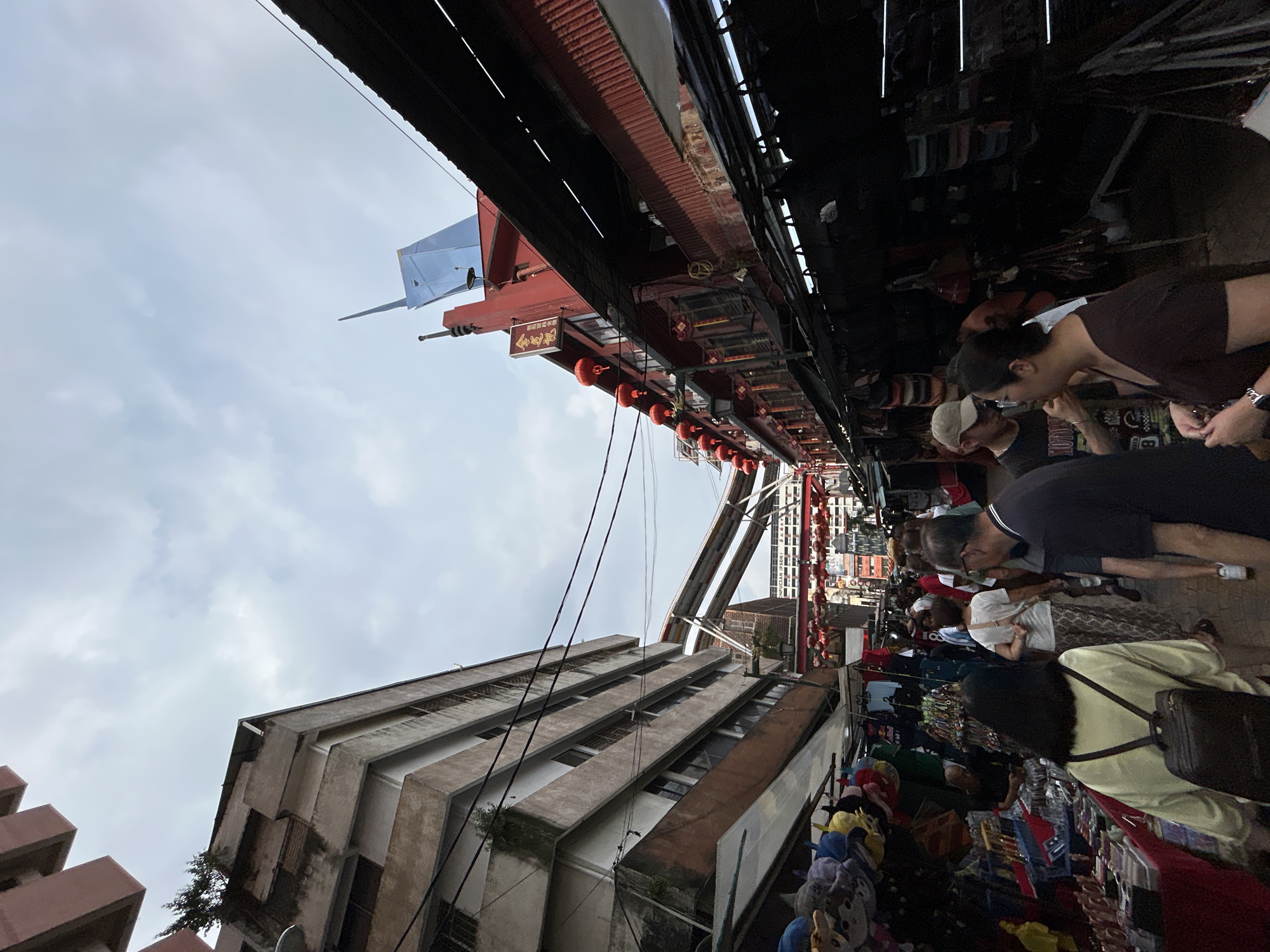 This eye-level shot captures a street scene in Kuala Lumpur, Malaysia. The image, oriented diagonally, features pedestrians browsing merchandise along a crowded street. The architecture includes a multi-story building with prominent concrete ledges and an overhead structure decorated with red lanterns and signage in Chinese characters. A metallic, angled structure extends into the sky. The sky is overcast. Merchandise is displayed on tables and racks along the street. People are interacting by examining the wares. Overhead, an extensive canopy or roof structure provides shade. Electrical wires intersect the scene.