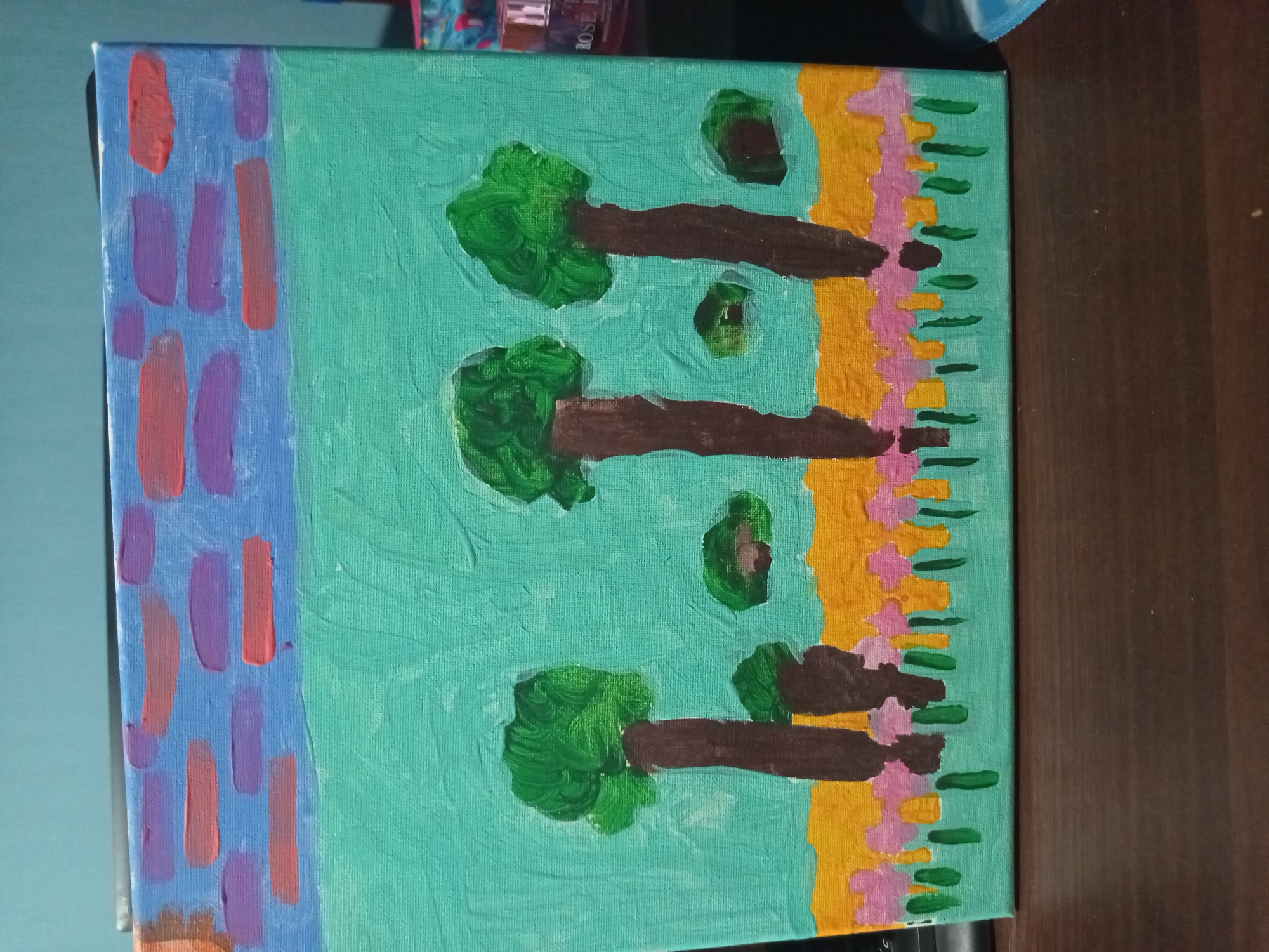 A flat canvas painting depicts a stylized landscape. The sky is a light blue with horizontal strokes of purple and coral, resembling abstract clouds. Below the sky is a solid band of turquoise, representing a field. In this field, three tall, brown-trunked trees with dense green foliage are spaced unevenly. Two smaller, green clumps of vegetation are also present in the field. A band of yellow runs along the bottom of the turquoise field, and below that, a line of small pink flowers with green stems is painted. The painting is placed on a dark wooden surface, possibly a desk or table. A keyboard is partially visible on the left side of the frame, and a cylindrical object, possibly a bottle, with a label reading "ROSE" is visible on the right. The background is a plain blue wall. The image is a photograph taken from a slightly elevated angle, looking down at the painting.