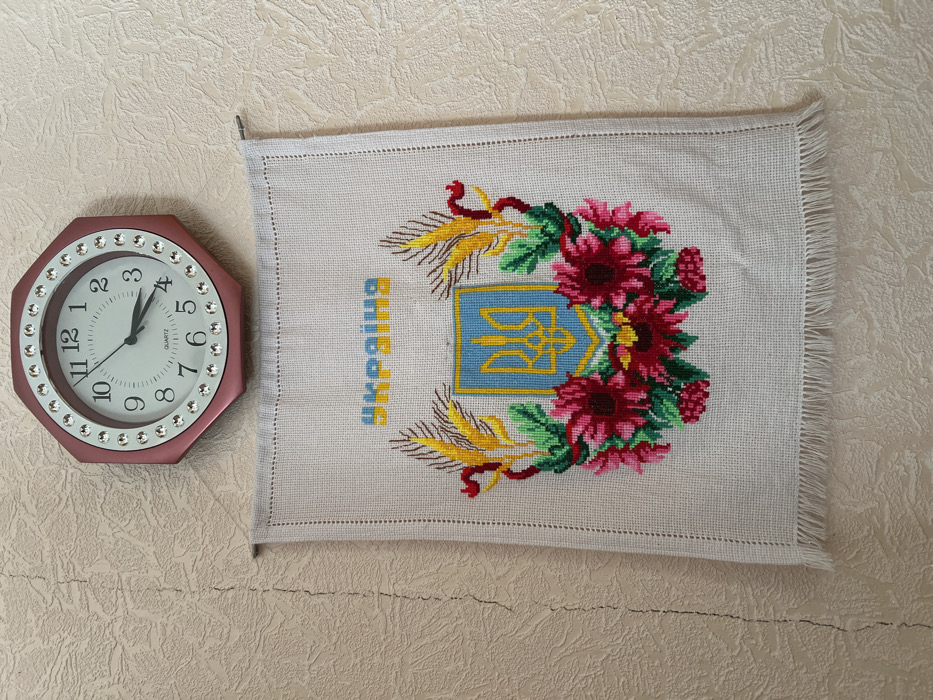 The scene depicts an interior wall in Pyriatyn, Ukraine. The wall is covered in light beige or off-white textured wallpaper, exhibiting a vertical crack on the left side. Two items are mounted on the wall. In the upper center, an octagonal wall clock with a pinkish-red frame is present, featuring clear, round decorative jewels around its perimeter. The clock has a white face with black Arabic numerals from 1 to 12. The hour hand points approximately halfway between 3 and 4, and the minute hand points directly at 4, indicating a time of 3:20. The word "QUARTZ" is printed below the center of the clock face. Directly beneath the clock, an embroidered textile hangs from a thin metal rod. The fabric is a light, natural color, possibly linen or cotton. The embroidery features a central Ukrainian national emblem, the Tryzub (trident), rendered in yellow on a blue shield with a yellow outline. This shield is flanked by two stylized golden-yellow wheat stalks with brown accents and is surrounded beneath by a semi-circular wreath of deep red multi-petaled flowers with dark centers, intermingled with green leaves and smaller red berry-like clusters. Above the emblem, the word "УКРАЇНА" (Ukraine) is embroidered in yellow letters with blue outlines. The bottom edge of the textile features a fringe of loose threads.