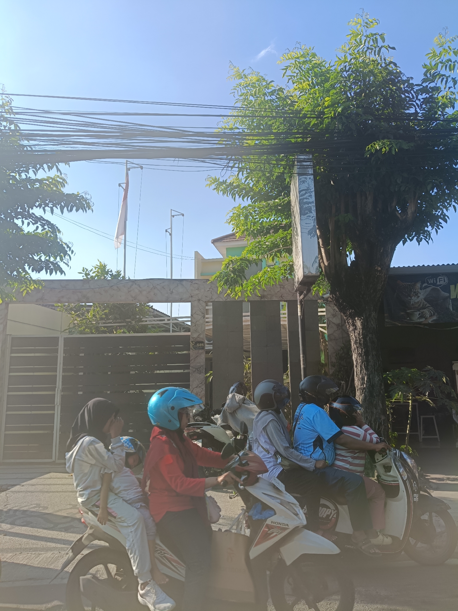 A group of people are riding on motorcycles in Klangonan, Indonesia. The scene depicts a sunny day with a clear blue sky. In the foreground, a motorcycle carries a woman wearing a red jacket and blue helmet, a young girl in a white outfit wearing a small blue helmet, and a child sitting in front of the woman. Behind them, another motorcycle is occupied by three people, including a person in a blue shirt and another in a striped shirt. The motorcycles are parked on a paved road in front of a gated property with a modern fence and a building behind it. Trees with lush green foliage are visible on the right side of the image, and power lines are strung across the top of the frame. A sign featuring a cat and "Wi-Fi" is mounted on the building. The overall impression is a snapshot of daily life and transportation in this Indonesian locale.