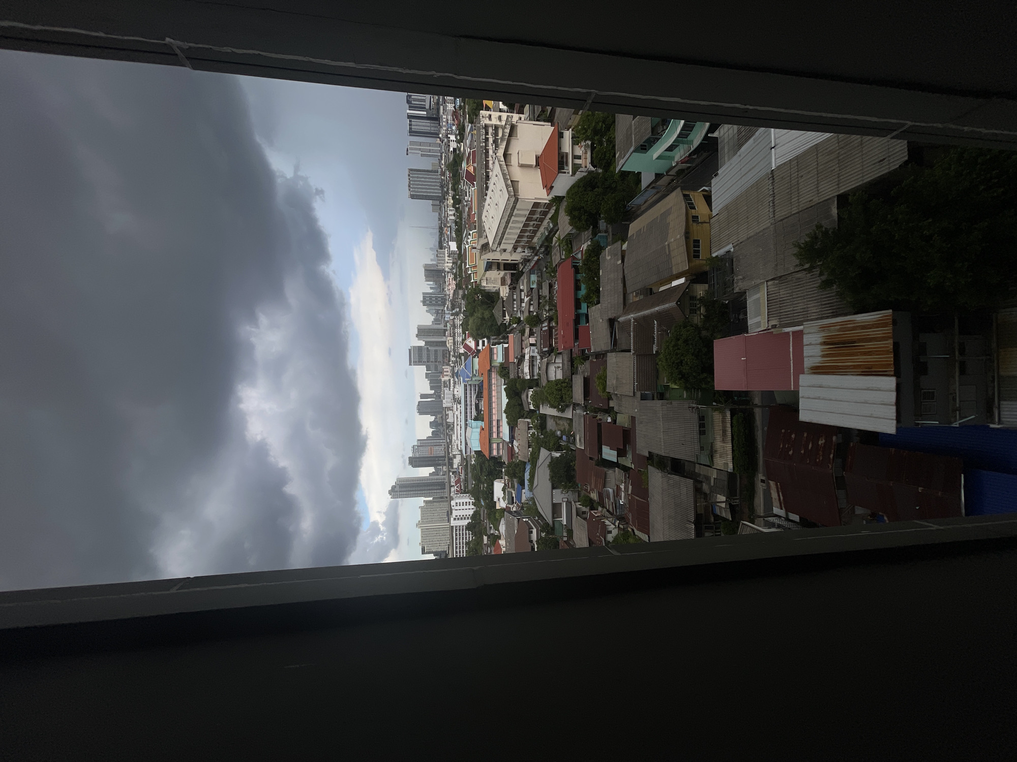 This media file displays an aerial view of a dense urban landscape, likely Bangkok, Thailand, as suggested by the location context. The scene is dominated by a mix of low-rise buildings with corrugated metal roofs and taller, more modern high-rise structures in the distance. The perspective is from an elevated vantage point, possibly a window, looking out over the city.
The weather appears to be overcast with dark, heavy clouds on the left side of the image, hinting at an impending storm or recent rainfall. Patches of brighter sky and clouds are visible to the right. The time of day is unclear due to the weather conditions, but it seems to be daylight.
The buildings exhibit a variety of architectural styles and colors, with many appearing to be residential or commercial structures. Some of the low-rise buildings seem to be packed closely together, indicating a densely populated area. The presence of trees interspersed among the buildings suggests pockets of greenery within the urban sprawl.
There are no people or explicit activities taking place that are discernible from the image. The focus is on the cityscape itself, presenting a view of urban development and its environmental context. The overall mood is somewhat somber due to the dark clouds, but the vibrant colors of some of the buildings offer a contrasting element. No text is visible in the image.