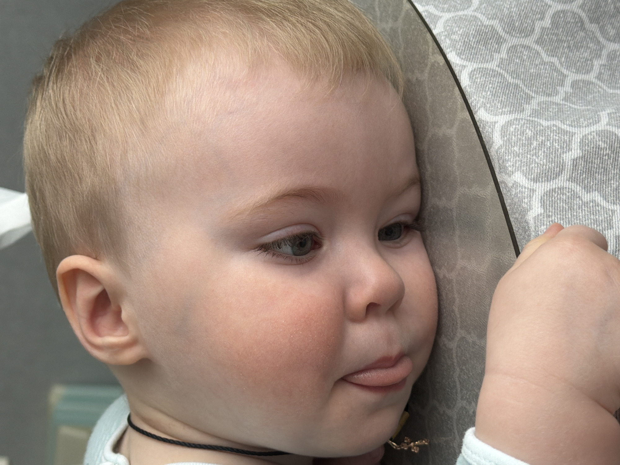 A close-up image depicts a young child, consistent with an infant or toddler, lying on their left side with their head resting on a grey, patterned surface. The child possesses fair skin, light blonde hair, and visible blue eyes. The left cheek is pressed against the resting surface, exhibiting a slight reddish tint. The left eye is partially open, directed downwards, and the tongue is observed slightly protruding from the mouth. A thin black cord encircles the child's neck, from which a small, gold-toned cross pendant hangs. The child's left hand, characterized by dimpled skin, rests on the same patterned surface in the lower left foreground. The surface is a textile material with a repeating light grey geometric pattern against a darker grey base. A distinct, thin dark line traverses horizontally across the middle of the frame. The background is out of focus and appears to be a light, indeterminate interior space. The specific location within Votkinsk, Russia, cannot be determined from the visual information provided.