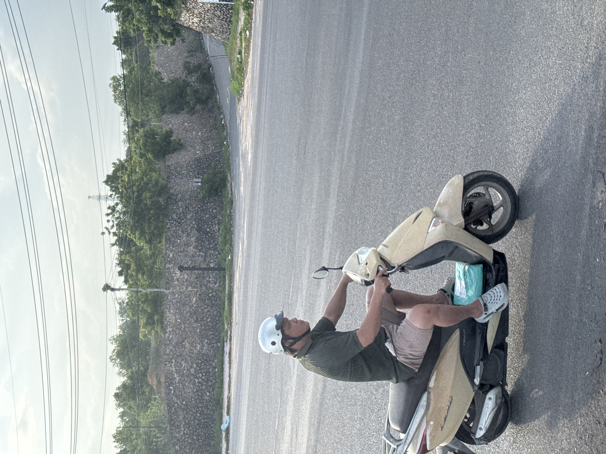 A man wearing a helmet and a green short-sleeved collared shirt rides a light-yellow scooter on an asphalt road in Mũi Né, Vietnam. He has on beige shorts and white shoes. On the scooter's footboard, a bright turquoise package is visible. The background features a low stone wall, lush green trees, and utility lines against a hazy sky.