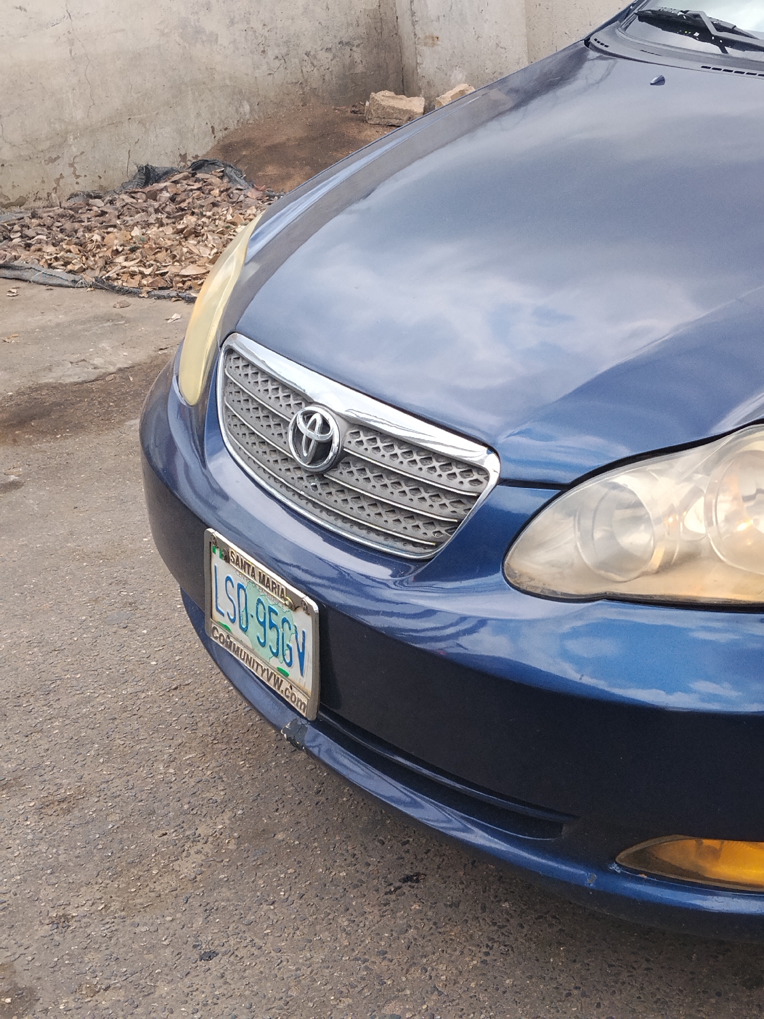 The image captures the front-right portion of a blue Toyota Corolla car, likely parked outdoors during daytime under an overcast sky. The car's metallic blue paint reflects the sky, showing soft, diffuse lighting. The main subjects are the car itself and its immediate surroundings.
The vehicle is an older model Toyota Corolla, identifiable by its grille design and the prominent Toyota emblem. Its front-right headlight appears yellowed and oxidized, a common sign of aging. Below the grille, the license plate frame is visible, bearing the text "SANTA MARIA" at the top and "COMMUNITYVW.com" at the bottom. The actual license plate displays the alphanumeric sequence "LSD-956GV" in green text on a white background.
The car is situated on a gray, coarse asphalt or paved surface, which shows some dirt and minor imperfections. To the left of the car, against a light-colored, rough concrete wall, is a pile of dried, brownish leaves or wood chips resting on a dark mesh or tarp. Some loose concrete blocks are also visible near the wall. The overall scene suggests an urban or semi-urban outdoor setting, consistent with Ikeja, Nigeria. No people or active events are depicted.