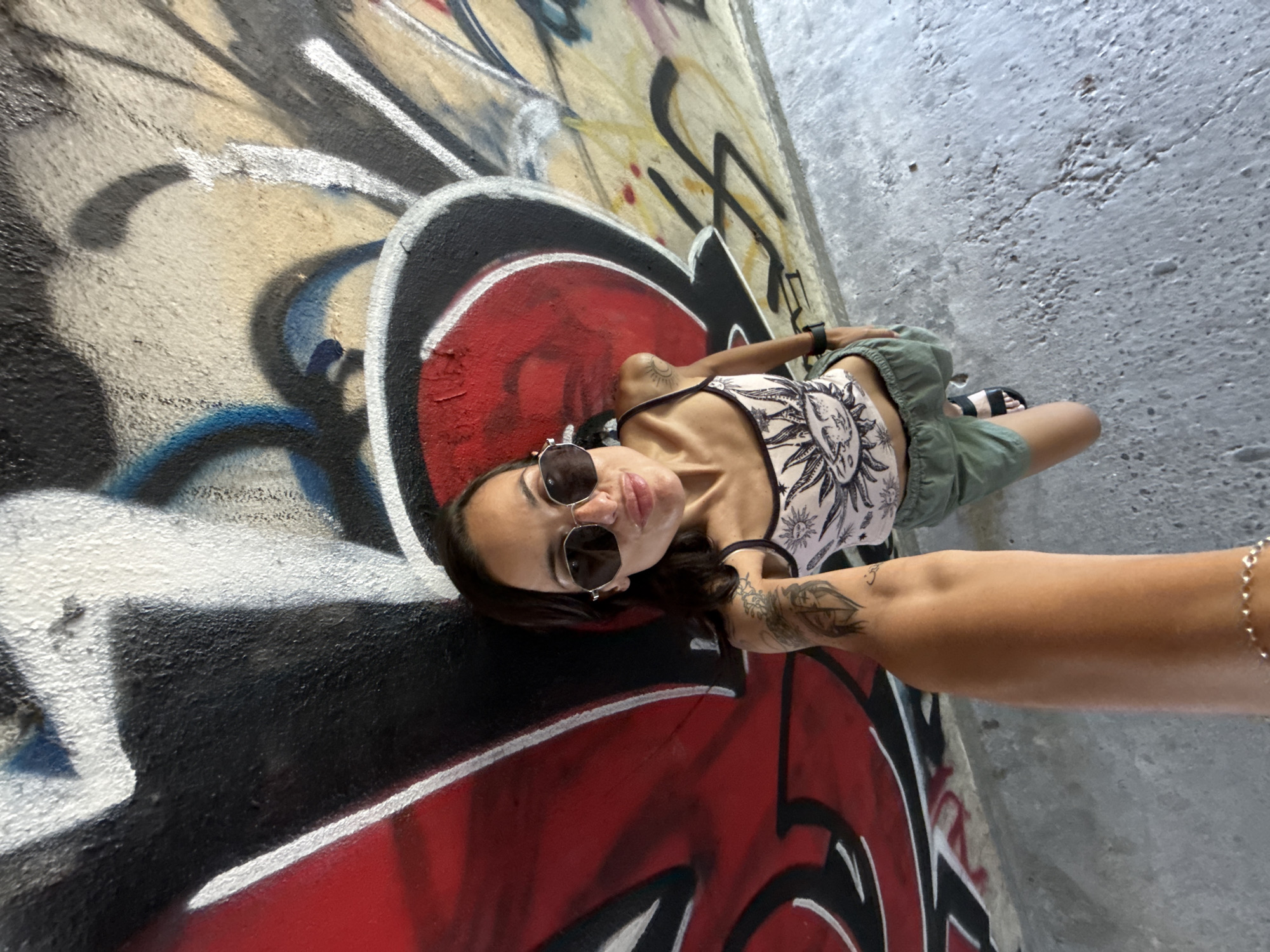 A high-angle, full-body selfie is taken by a woman standing in front of a graffiti-covered wall. The woman has dark hair, is wearing sunglasses, a tank top with a sun design, and green shorts. She is holding the camera out with her left arm, and her right arm is visible in the foreground of the image. The graffiti on the wall is colorful and abstract, with large black, red, and white spray-painted shapes. The ground beneath her is concrete. The location is identified as the underpass of the Pleven Railway Station in Pleven, Bulgaria.