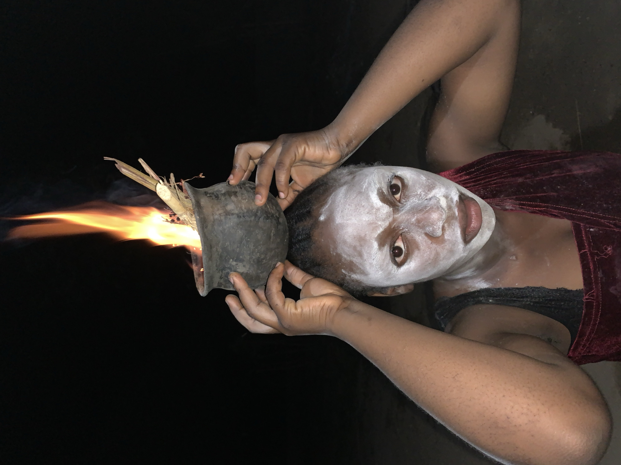 A person with dark skin, covered in white powder on their face, is holding a dark, primitive pot that contains a fire with flames emanating from it. The person is wearing a dark-colored top and a maroon shawl or wrap. The background is completely black. There is no indication of a location in Kukum, Nigeria or a city environment.