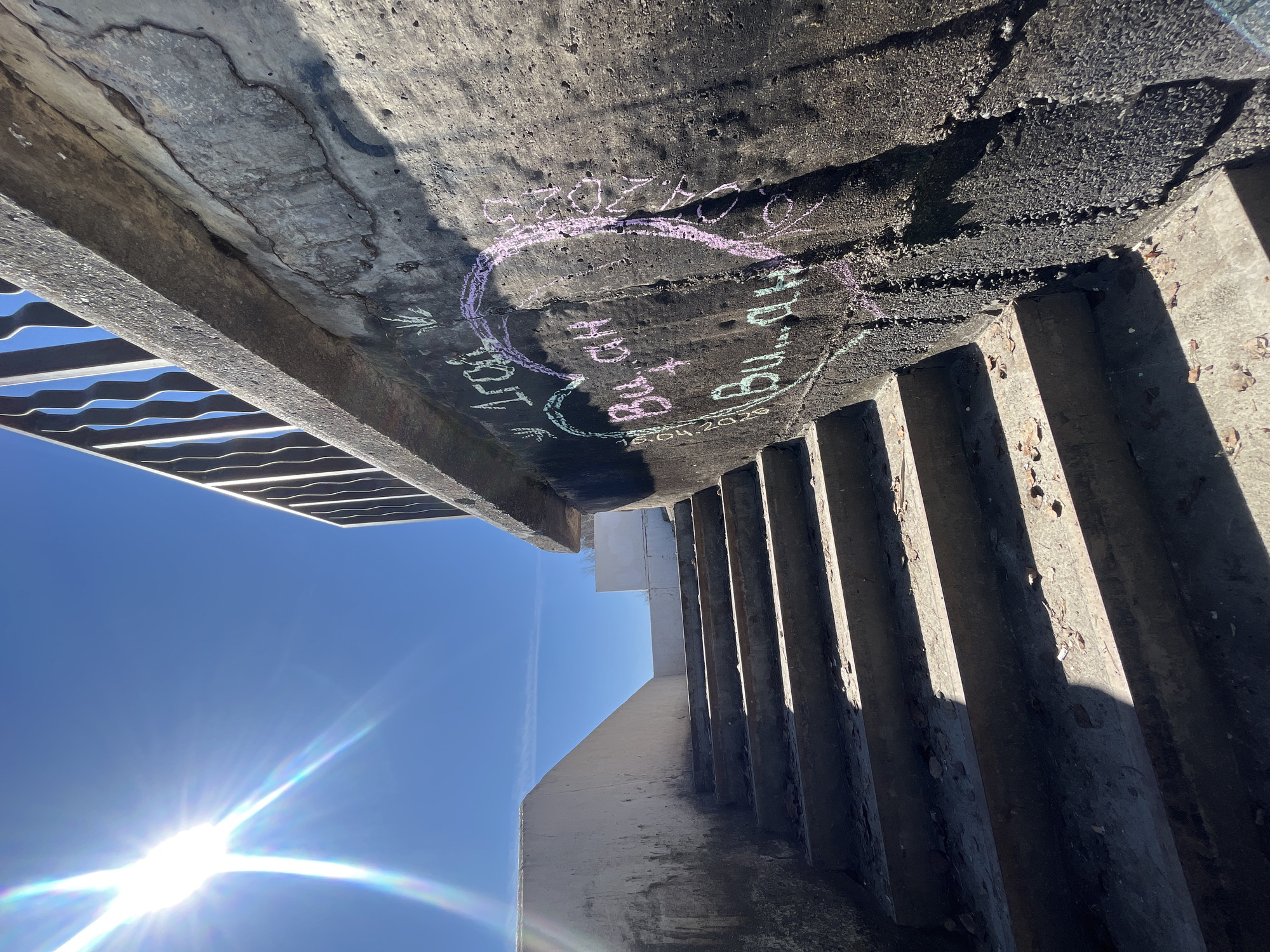 A low-angle shot from beneath a concrete structure captures sunlight and architectural details. In the sky, a bright sun creates a glare, illuminating the clear blue. The structure's underside is textured, marked with graffiti including names and dates (e.g., "Szczotka", "HD + Bu" and "Bu + BU, 16.04.2023") written in chalk, along with a drawing of a heart. To the left, a metal grate suggests a walkway or stairs, while on the right, concrete supports form a row of vertical lines casting shadows. The location is Chaikovsky, Russia, based on the graffiti.