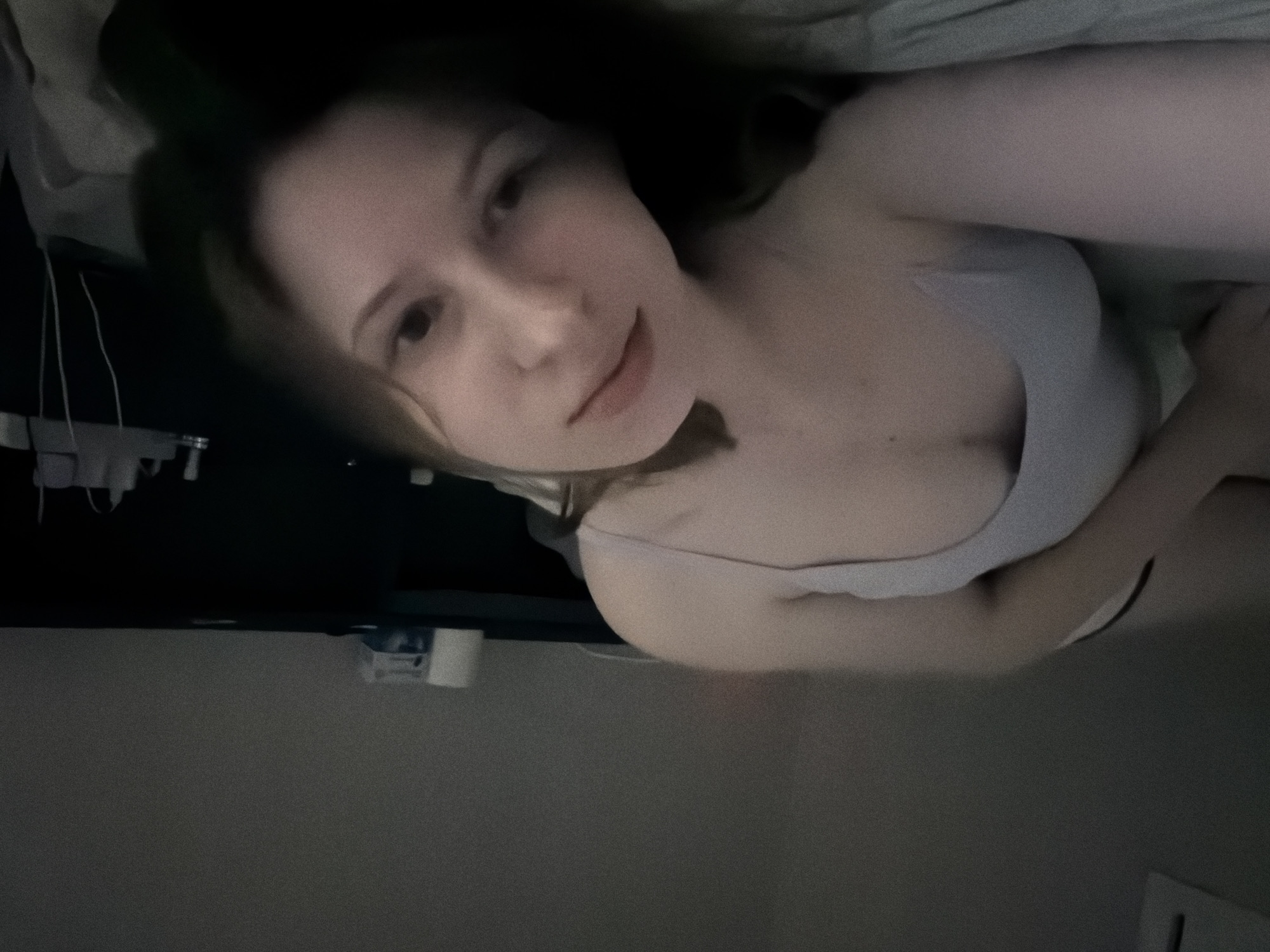 The image shows a person lying on a bed, facing the camera. The person is wearing a light-colored tank top and underwear. Their body is positioned on its side, with their head turned towards the viewer. The background appears to be a dimly lit room, possibly a bedroom, with a dark surface or furniture visible behind the person. There are some objects on this surface, including what appears to be a roll of toilet paper and some packaging. The overall lighting is low, creating shadows and a somewhat grainy texture to the image. The person's facial features are visible, and they are looking directly at the camera with a neutral expression. The location is not identifiable from the visual information in the image.