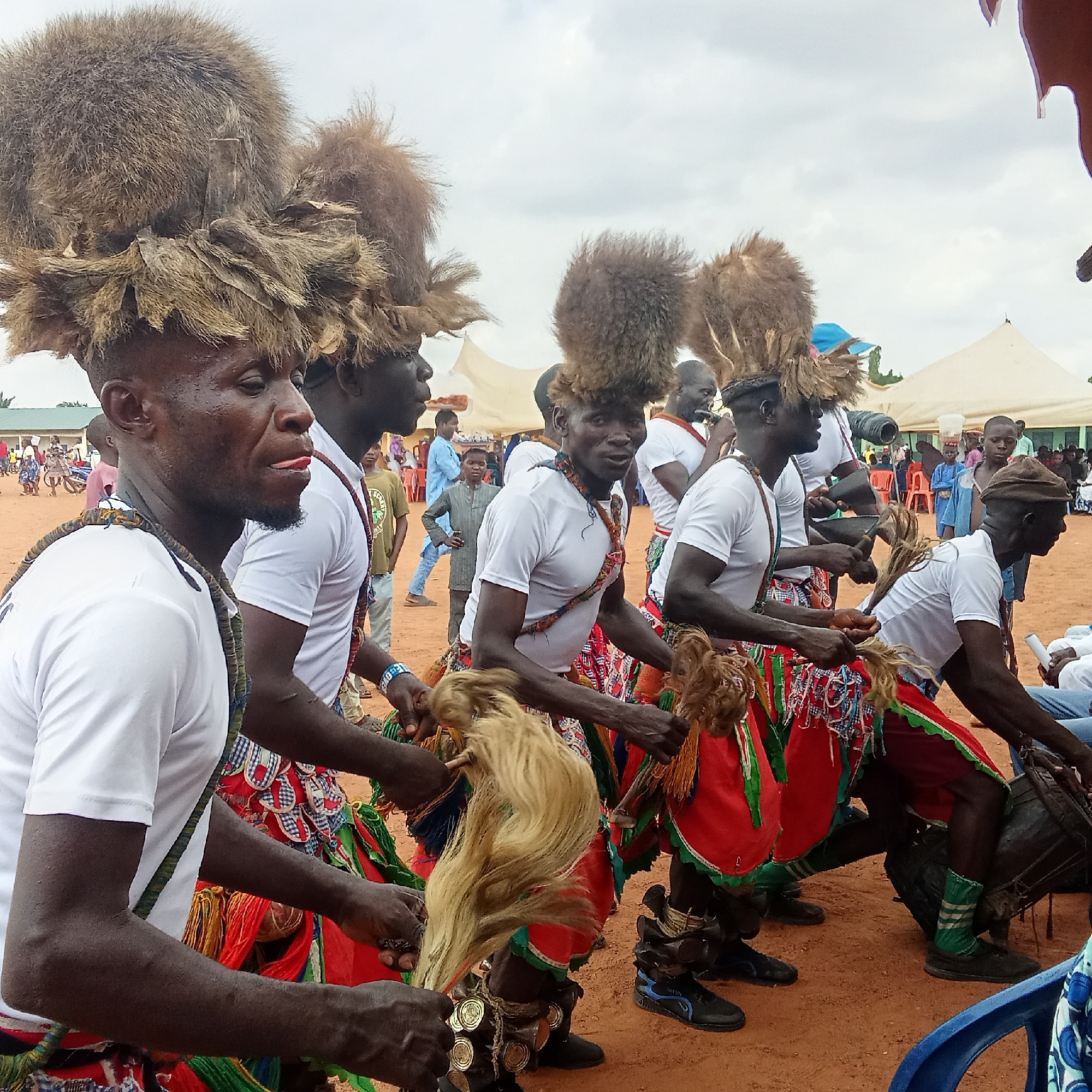 A group of men in Akwanga, Nigeria perform a cultural dance. The men wear matching white t-shirts, decorative skirts of red, green, white and black, and large furry head coverings. They carry blonde, straw-like items in their hands and wear noise-making decorations on their ankles. At the end of the line, one of the men plays a drum. The performance takes place on a red, sandy ground with onlookers of all ages in the background and white tents in the distance. The sky is grey and overcast.