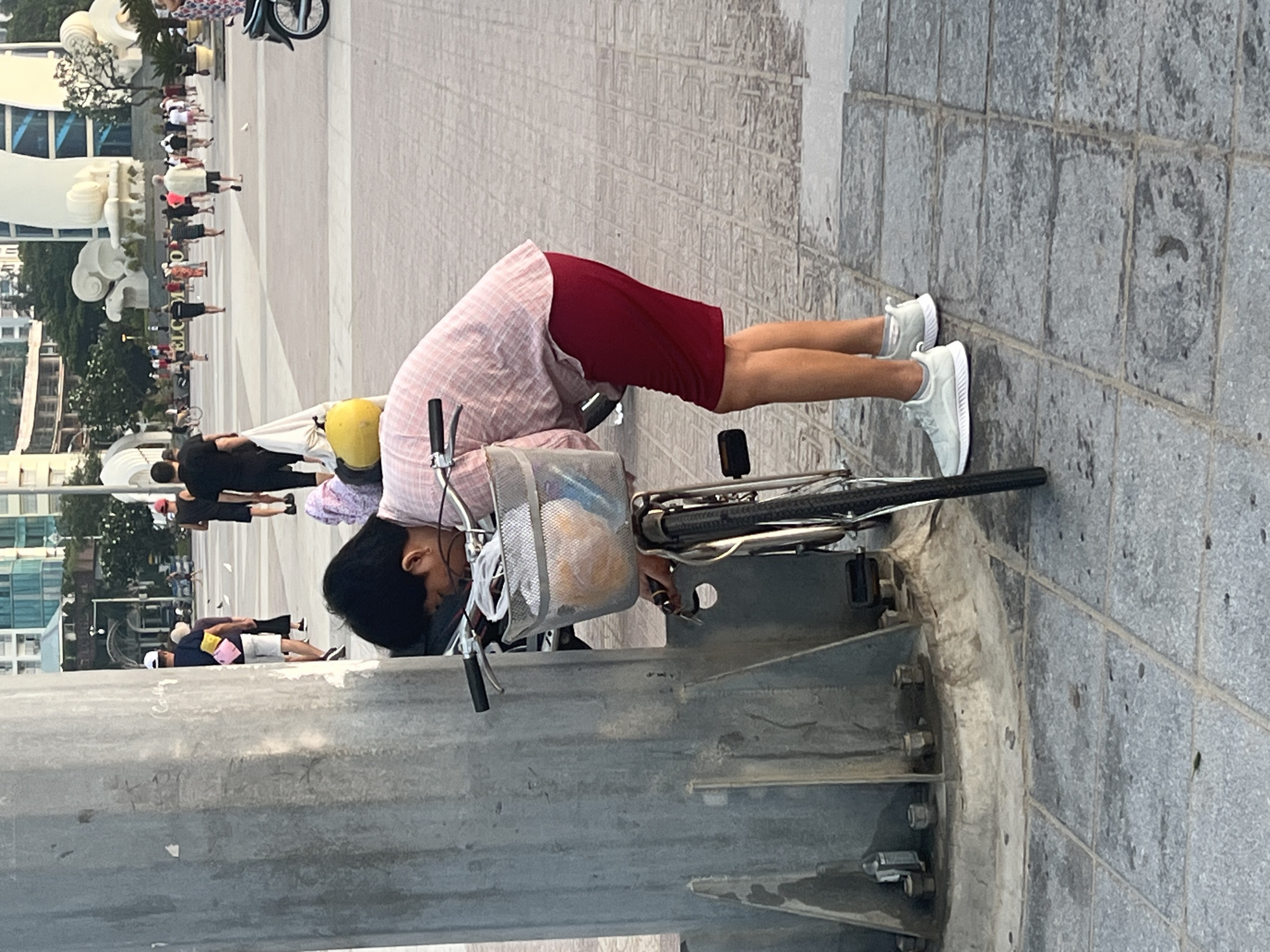 A woman wearing a pink and white patterned shirt and a red skirt is bent over, seemingly working on a bicycle that is leaning against a grey concrete post. The bicycle has a wire basket on the front. The woman is wearing white sneakers. In the background, people are walking on a plaza, with buildings visible in the distance. This location is in the city of Nha Trang, Vietnam.