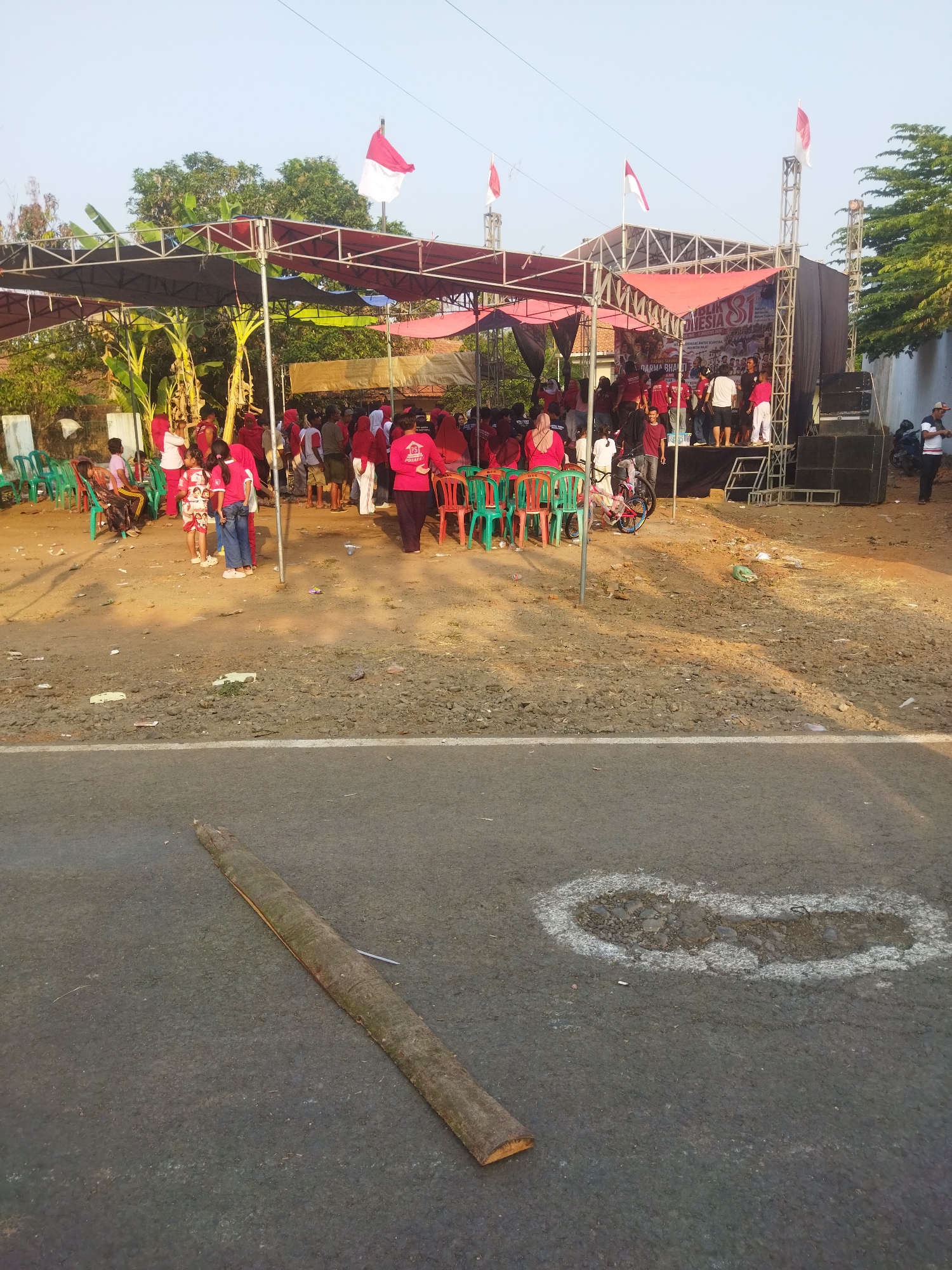 In the city of Banjar, Indonesia, an outdoor stage with a red canopy is set up for an event. Several flags, including the Indonesian flag, are visible. A crowd of people, many wearing red clothing, are gathered around the stage. Some are seated in green chairs, while others are standing. A bicycle is parked near the stage. The ground in front of the stage is unpaved and appears to be dirt and gravel. In the foreground, a road is visible with a fallen log and a chalk outline of a pothole. The scene suggests a community event or celebration taking place in a public space.